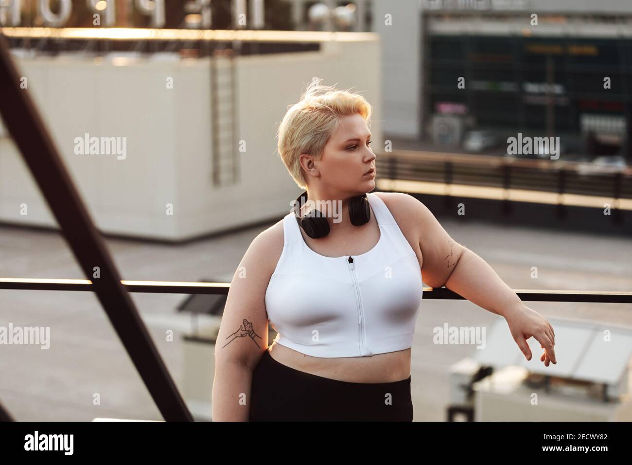 Frau mit übergroßem Körper, die sich während eines Trainings auf einem Dach an ein Geländer lehnt. Übergroße Fitness Frau in Sportbekleidung trägt Kopfhörer Blick weg outdoo Stockfoto
