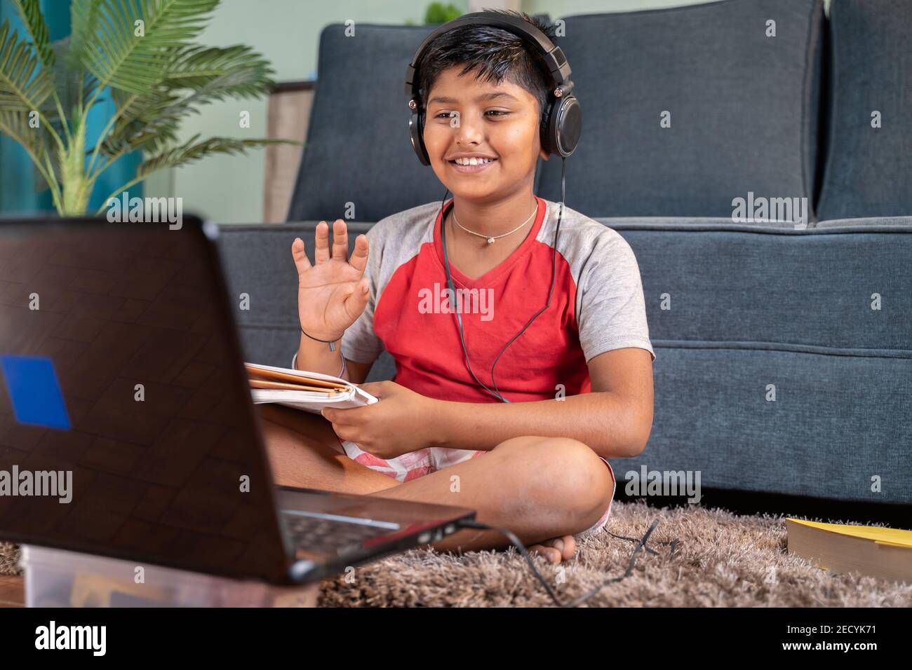 Kleines Kind mit Kopfhörern und Büchern im Handtelefon begrüßt seine Tutor während der Online-Klasse auf Laptop zu Hause - Konzept Des virtuellen Educatiom Stockfoto