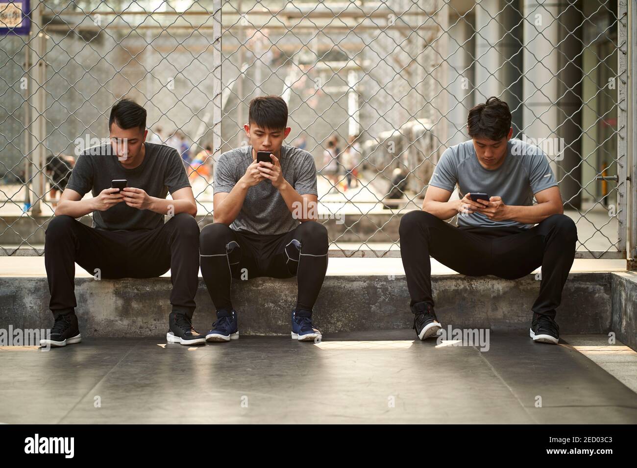 Drei junge asiatische Erwachsene Männer sitzen auf Boden Blick auf Mobiltelefon im Freien Stockfoto