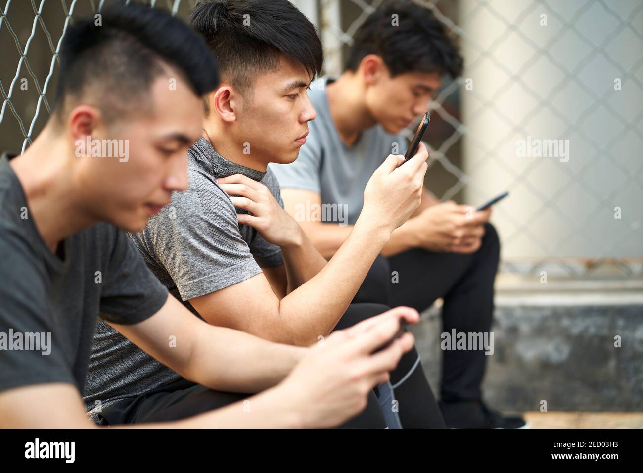 Drei junge asiatische Erwachsene Männer sitzen auf Boden Blick auf Mobiltelefon im Freien Stockfoto