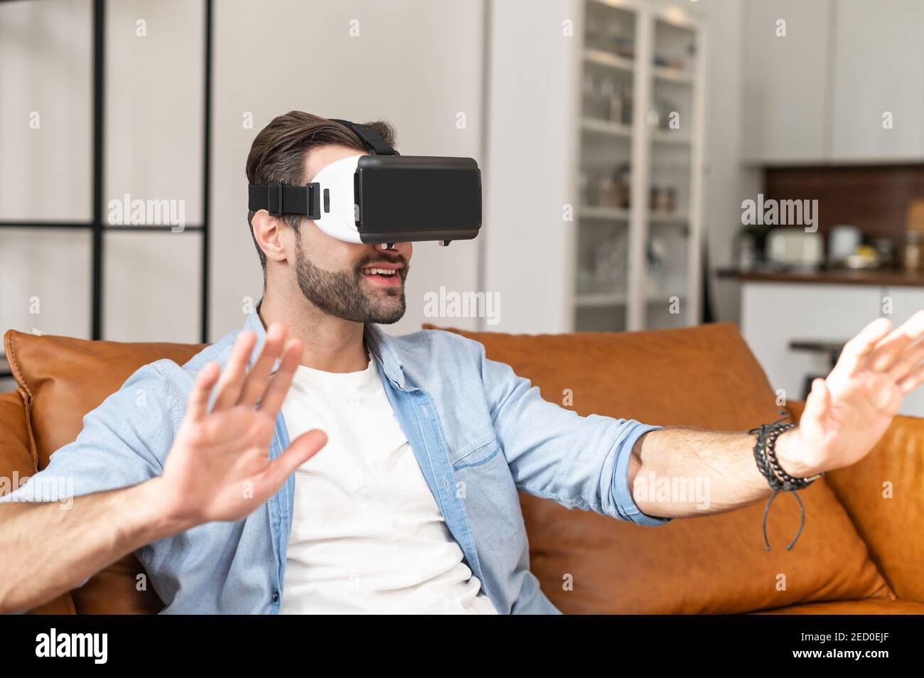 Aufgeregt junge bärtige Mann trägt Brille der virtuellen Realität sitzt auf der bequemen Couch zu Hause, ein Kerl in VR-Headset verloren im Cyberspace, mit Stockfoto