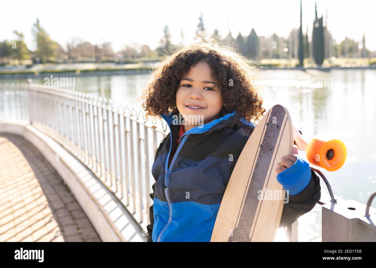 Entzücktes ethnisches Kind mit Afro-Frisur stehend mit Longboard auf Böschung an sonnigen Tagen und Blick auf die Kamera Stockfoto