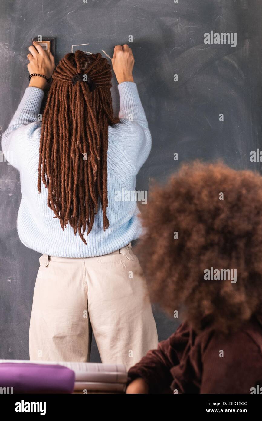 Rückansicht der Lehrerin in Dreadlocks Schreiben mathematisches Beispiel Auf Tafel für schwarzes Mädchen, das im Klassenzimmer am Schreibtisch sitzt Während des Unterrichts bei scho Stockfoto