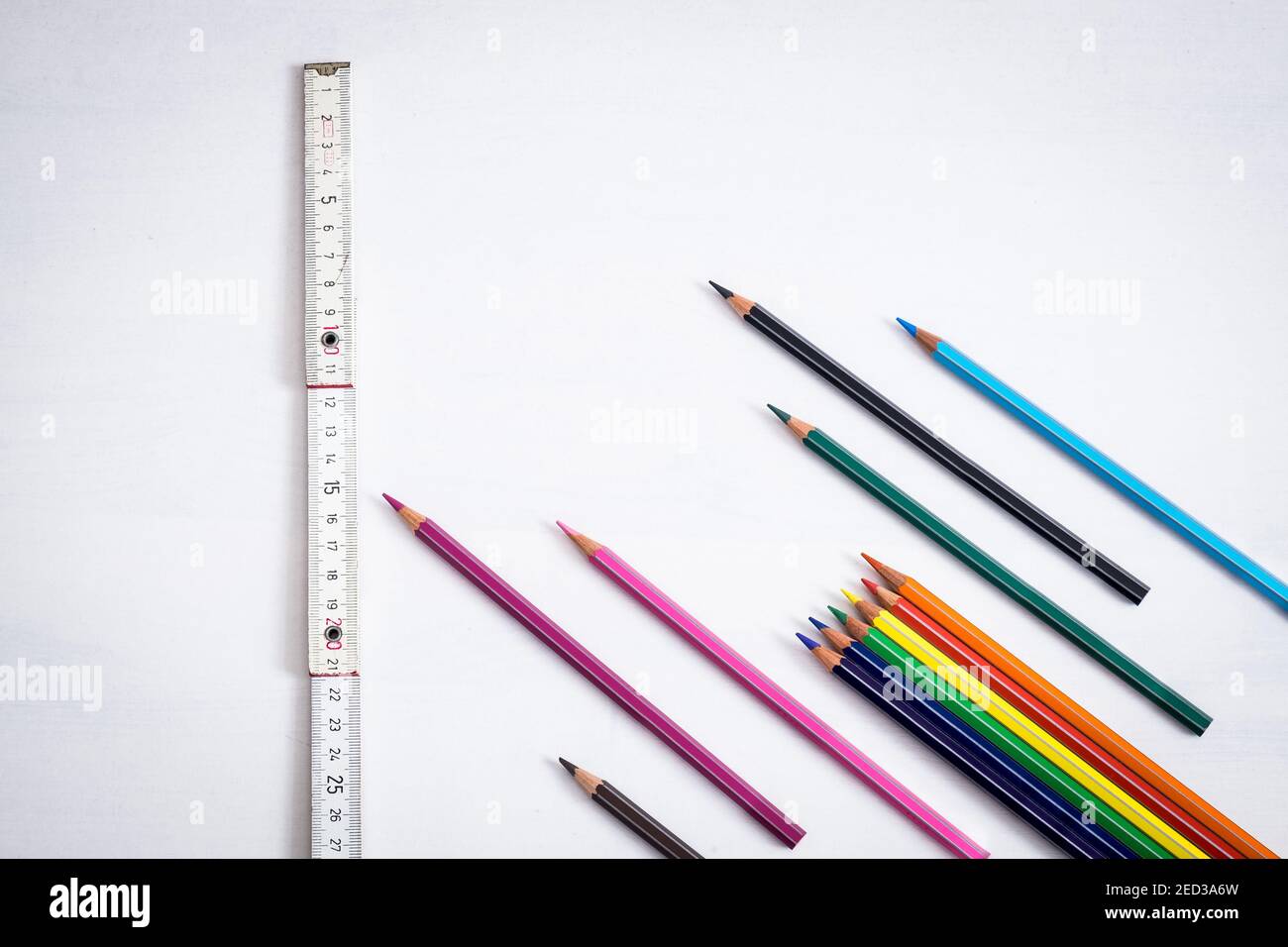 Viele farbige Buntstifte, die wie Pfeile auf einem weißen Hintergrund platziert sind, zeigt ein in der Nähe befindeglich Holzmesser den sicheren Abstand an, den man in den Klassenzimmern halten muss, um dies zu verhindern Stockfoto