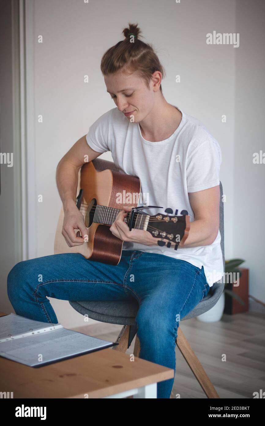 Echtes Lächeln eines jungen Sängers in einem weißen T-Shirt und Jeans sitzen auf einem Stuhl in der Mitte eines beleuchteten Raumes, spielen eine akustische Gitarre. Ein Mann mit b Stockfoto