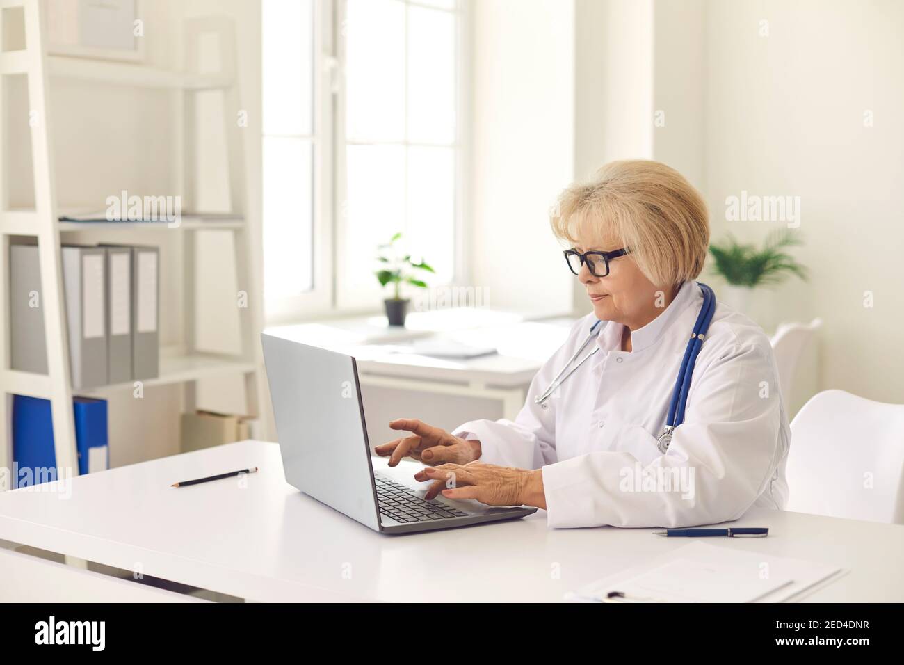 Reife Frau Arzt in weißen Uniform sitzen und arbeiten online Auf einem Laptop Stockfoto