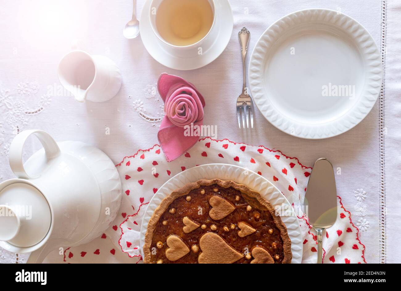 Ein Tisch für ein besonderes Frühstück. Eine orangefarbene Tarte auf einem Valentinsstoff wird mit einer Tasse Tee neben einer Serviette serviert, die wie eine Rose gefaltet ist. Stockfoto