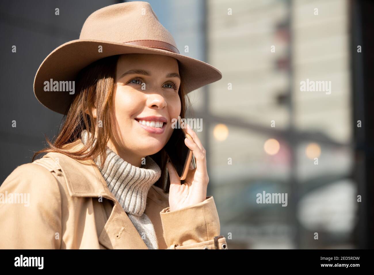 Happy Young wunderschöne Brünette weiblich in Hut, beige Trenchcoat und grauen Pullover im Gespräch auf Handy in städtischen Umgebung Stockfoto