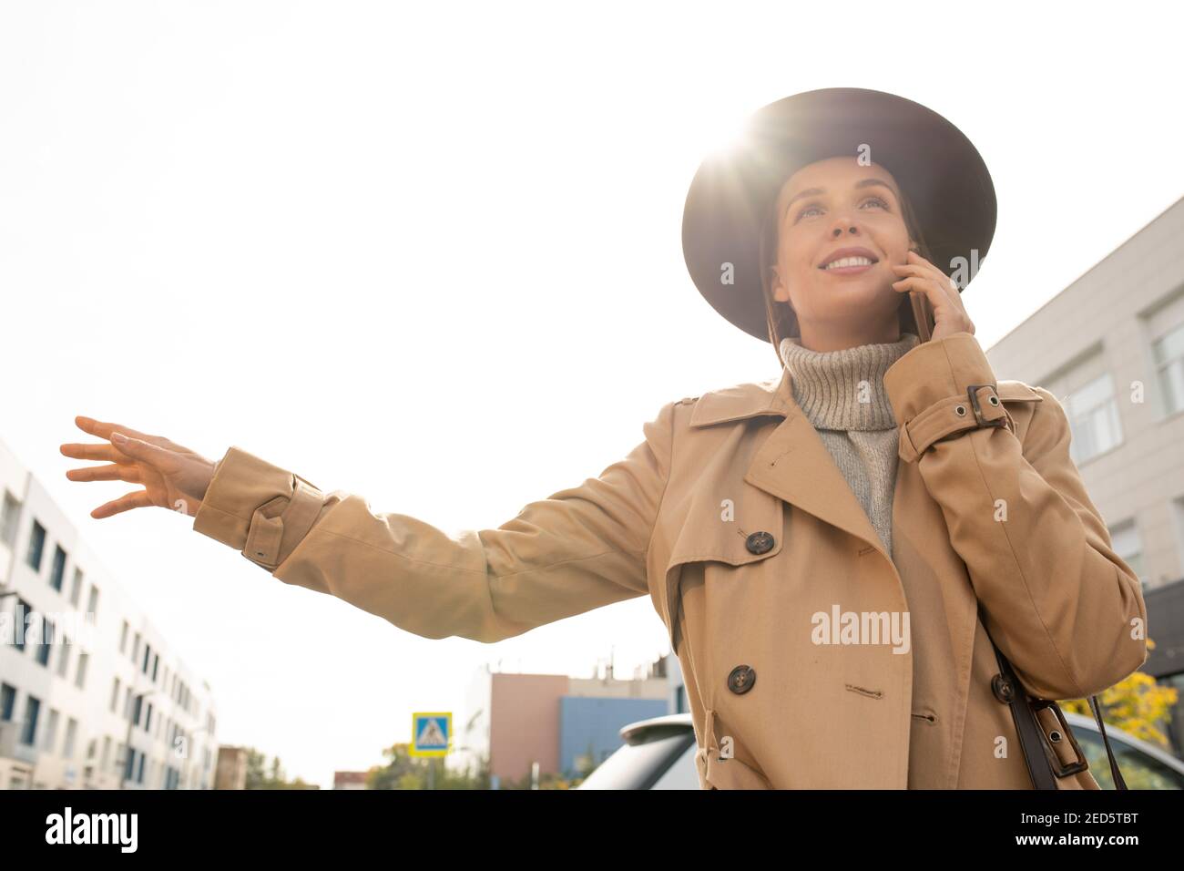 Junge elegante lächelnde Geschäftsfrau in beigem Trenchcoat und schwarzem Hut Versuchen, ein Taxi zu bekommen, während man auf der Straße in der Stadt steht Umwelt Stockfoto