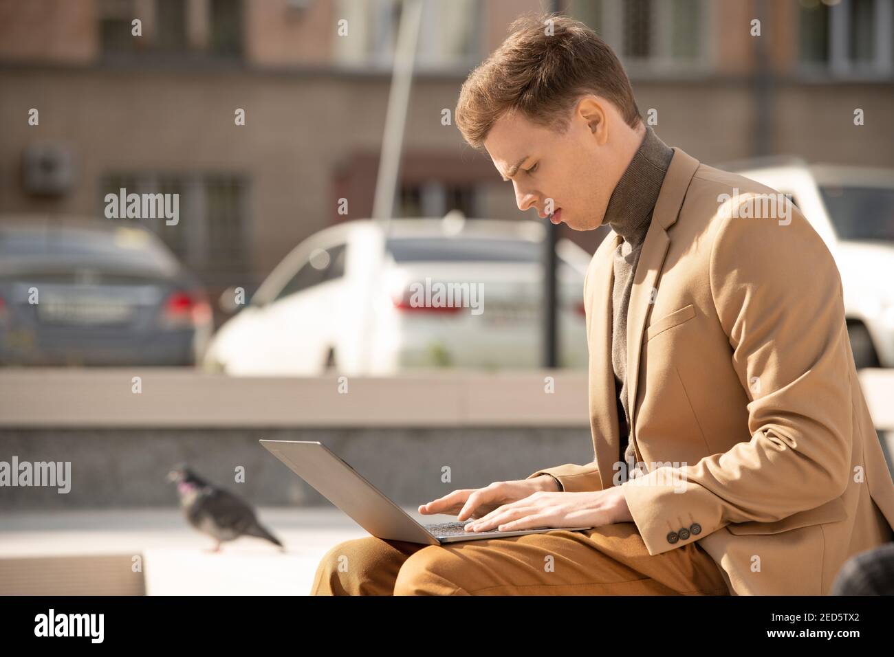 Junge seriöse Geschäftsmann in smart Casualwear sitzen in der städtischen Umgebung An sonnigen Tag und Surfen im Netz gegen Parkplatz Ort Stockfoto