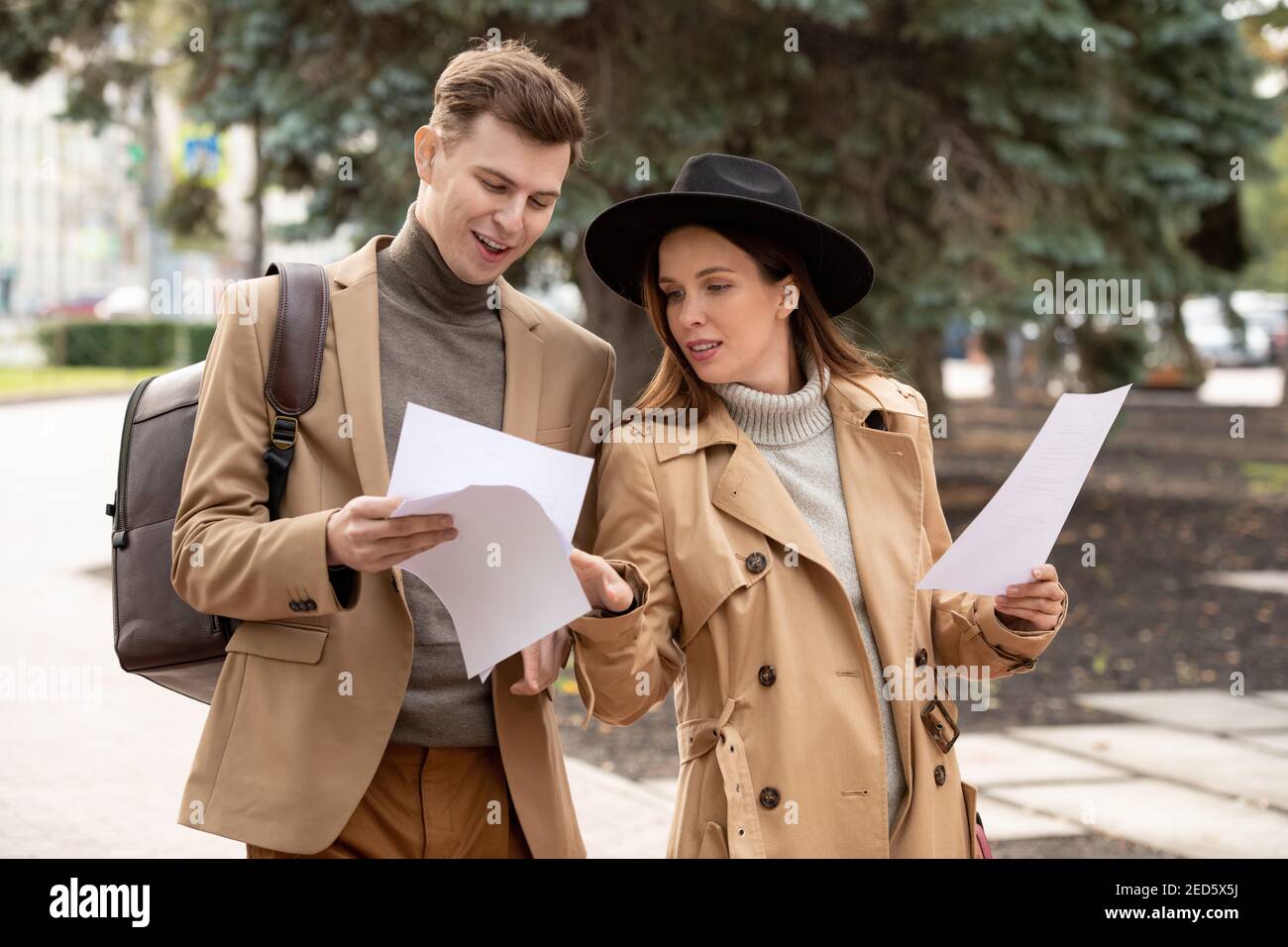 Zwei junge selbstbewusste Kollegen in Smart Casualwear lesen Papiere und Diskutieren sie während der Fahrt entlang der Straße im Park in der Pause Oder nach der Arbeit Stockfoto