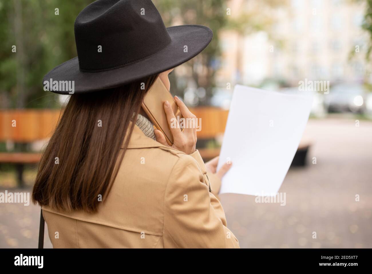 Rückansicht der jungen eleganten Geschäftsfrau in beigem Trenchcoat und Schwarzer Hut, der auf Papier schaut und auf dem Handy spricht Im städtischen Umfeld Stockfoto