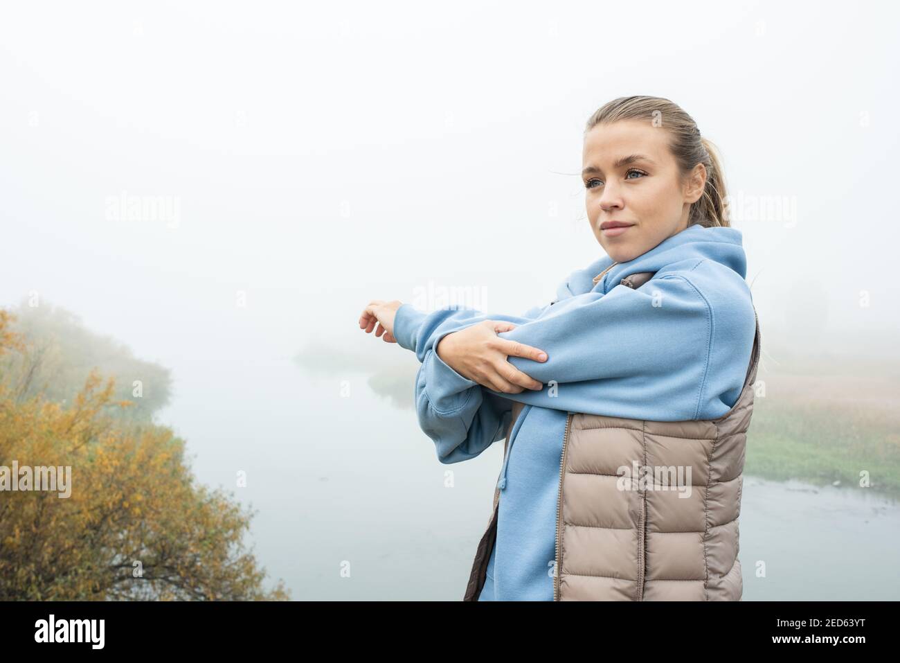 Young Fit Sportlerin in blauem Hoodie und grauem Jackentestung Ihr linker Arm vor sich selbst beim Sport hinein Natürliche Umgebung Stockfoto