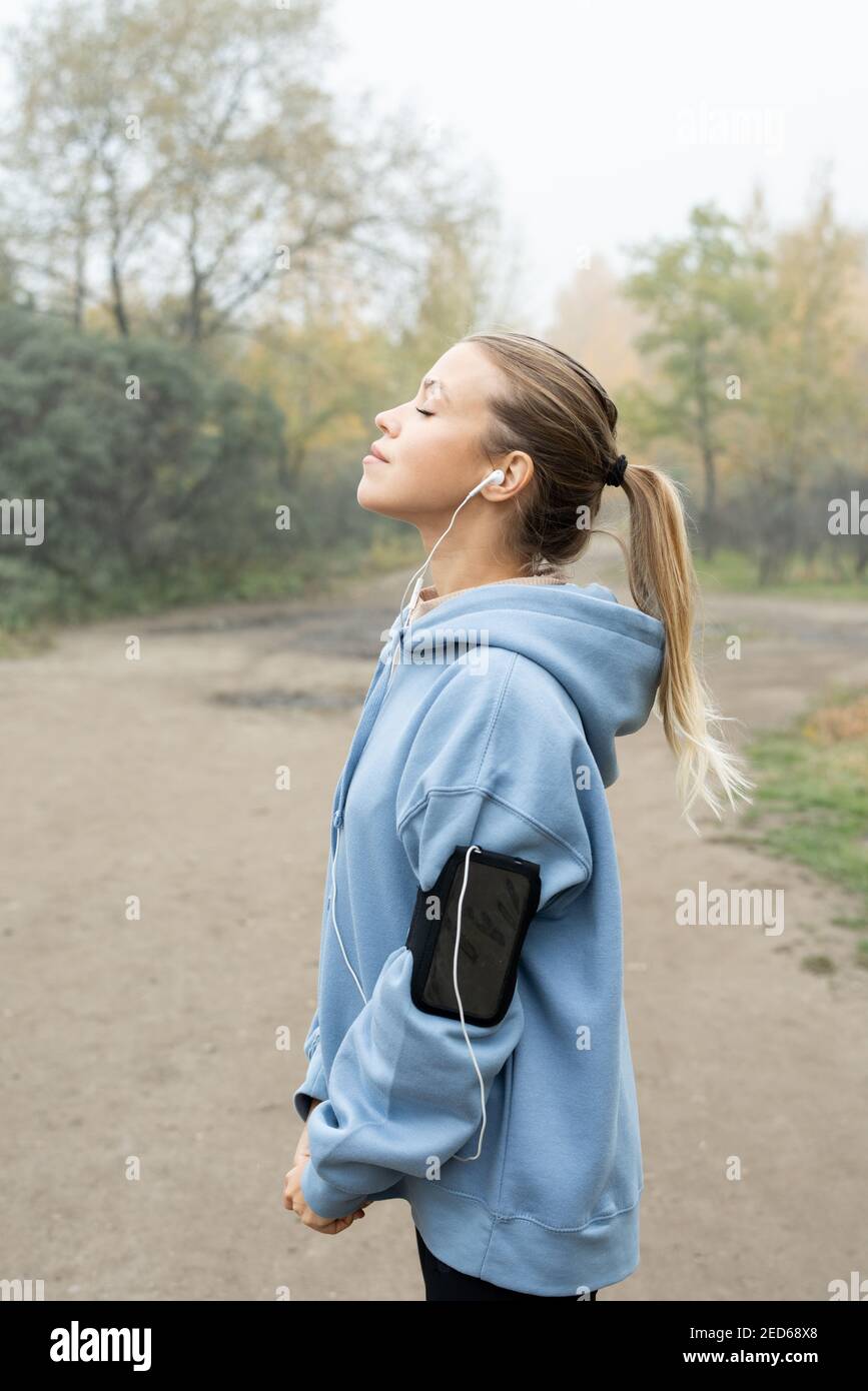 Seitenansicht der fröhlichen blonden Sportlerin mit Pferdeschwanz im Stehen Vorderseite der Kamera gegen Bäume und Straße im Park während Pause nach dem Joggen Stockfoto