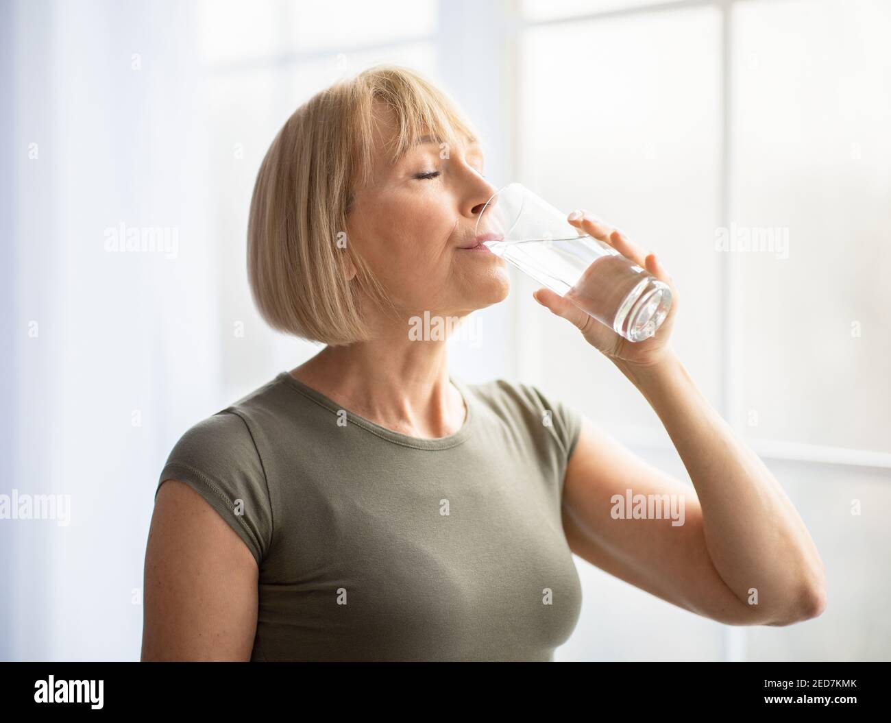 Fit ältere Frau trinken klares Wasser während ihrer Trainingspause zu Hause. Gesunder Lebensstil und Wellness-Konzept Stockfoto
