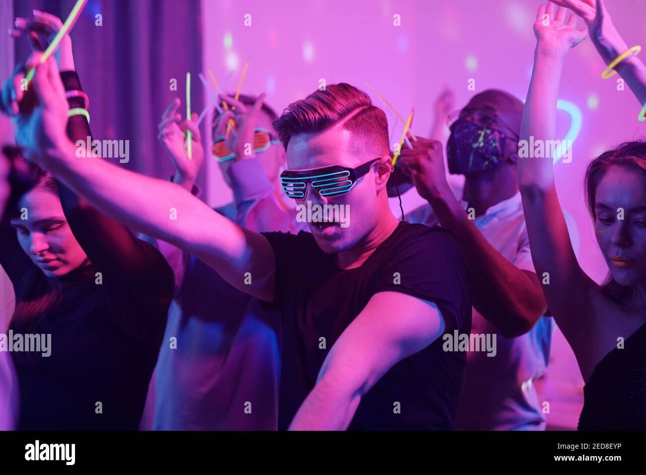 Junger aufgeregt Mann in Casualwear und Disco Brillen und seine Interkulturelle Freunde mit glühenden Stöcken tanzen zusammen, während sie zu Hause genießen Party Stockfoto