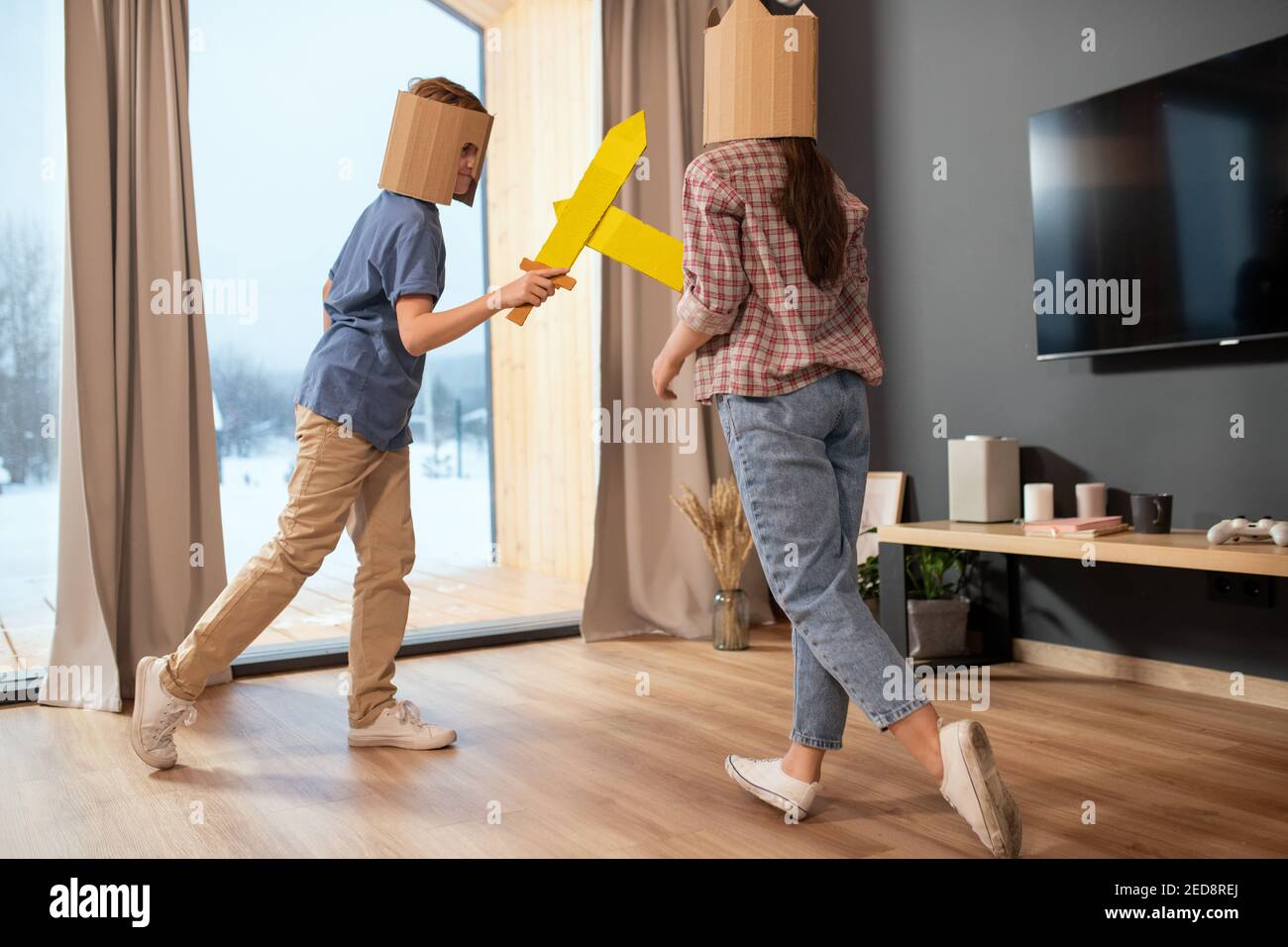 Zwei verspielte Geschwister in Casualwear und Papphelmen kämpfen mit Spielzeug Schwerter gegen große Fenster mit beigen Vorhängen im Wohnzimmer Stockfoto