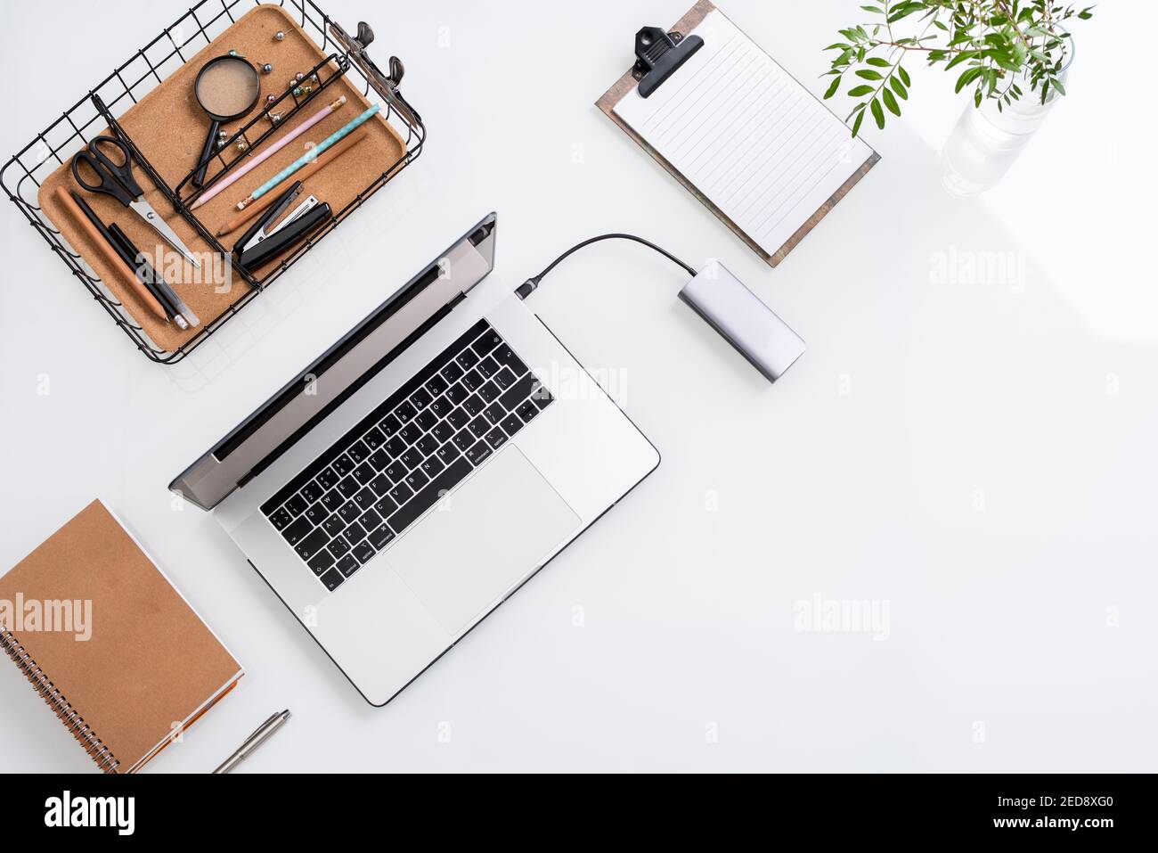 Draufsicht auf weißen Schreibtisch mit Laptop, Korb mit Bürobedarf, leeres Papier in Zwischenablage, grüne Pflanze in Glas, Notebook und silbernen Stift Stockfoto