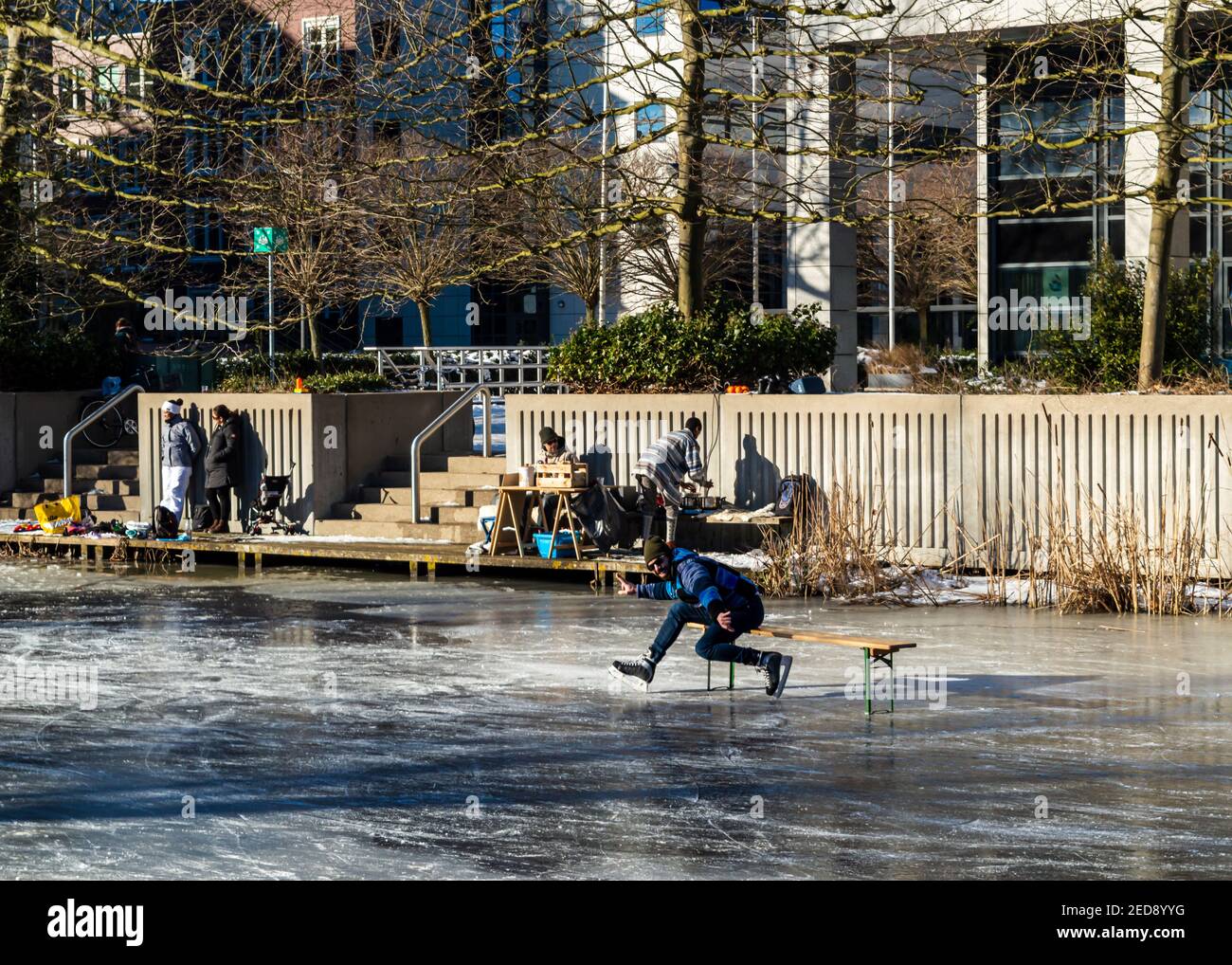 AMSTERDAM, NIEDERLANDE, FEBRUAR 13,2021. Eisläufer auf einer Holzbank auf dem gefrorenen Graben vor einem Gebäude, das eine lustige Pose und Menschen in t Stockfoto