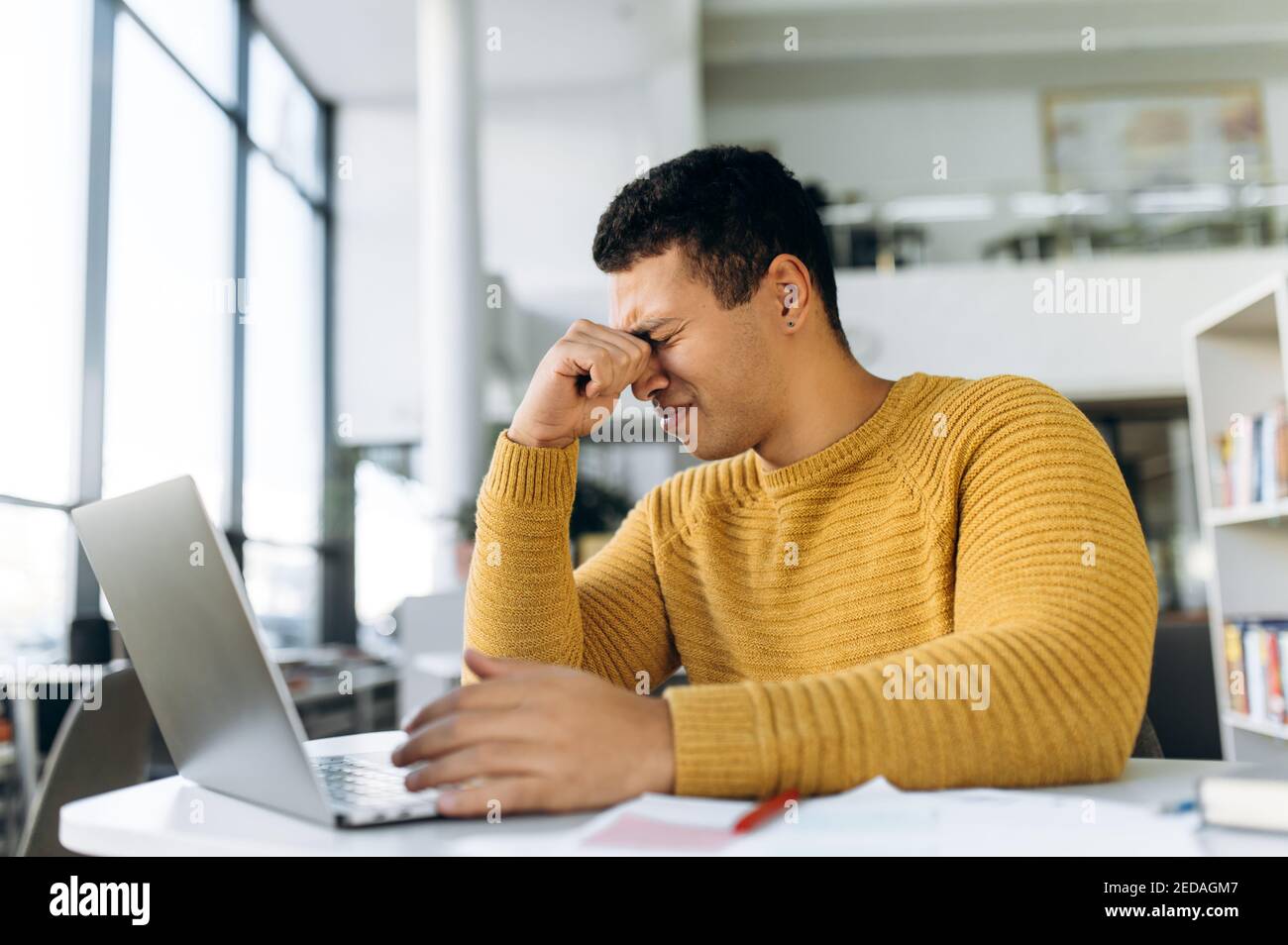 Erschöpfter männlicher Student oder Mitarbeiter, der am Tisch sitzt, massiert die Augen und hat Kopfschmerzen. Hispanischer Geschäftsmann müde von der Arbeit oder Studium, Gefühl ungesund oder gestresst Stockfoto