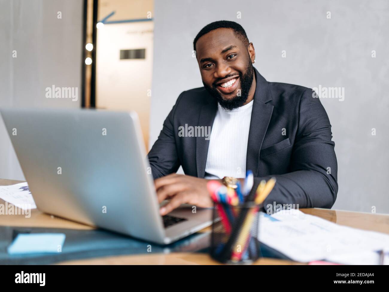 Erfolgreicher männlicher Manager oder Chef sitzt am Schreibtisch im Büro. afroamerikanischer Geschäftsmann mit Laptop, entwickeln neues Projekt oder Startup. Ein gutaussehender, selbstbewusster Mitarbeiter schaut lächelnd auf die Kamera Stockfoto