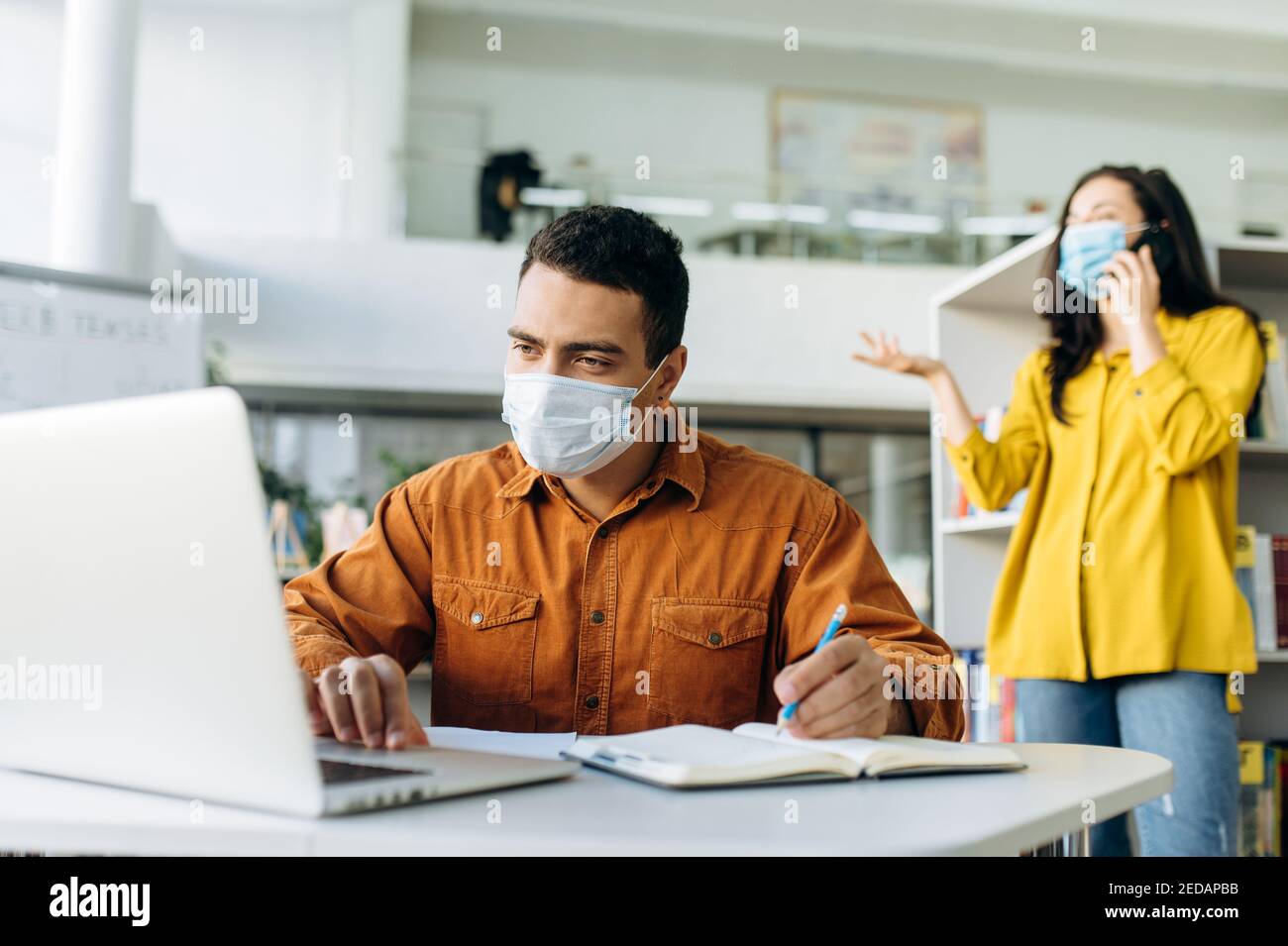 Erfolgreicher männlicher Manager oder Student mit Schutzmaske, arbeitet an einem Business-Projekt mit einem Laptop, im Hintergrund spricht ein Kollege am Telefon in einer Schutzmaske Stockfoto