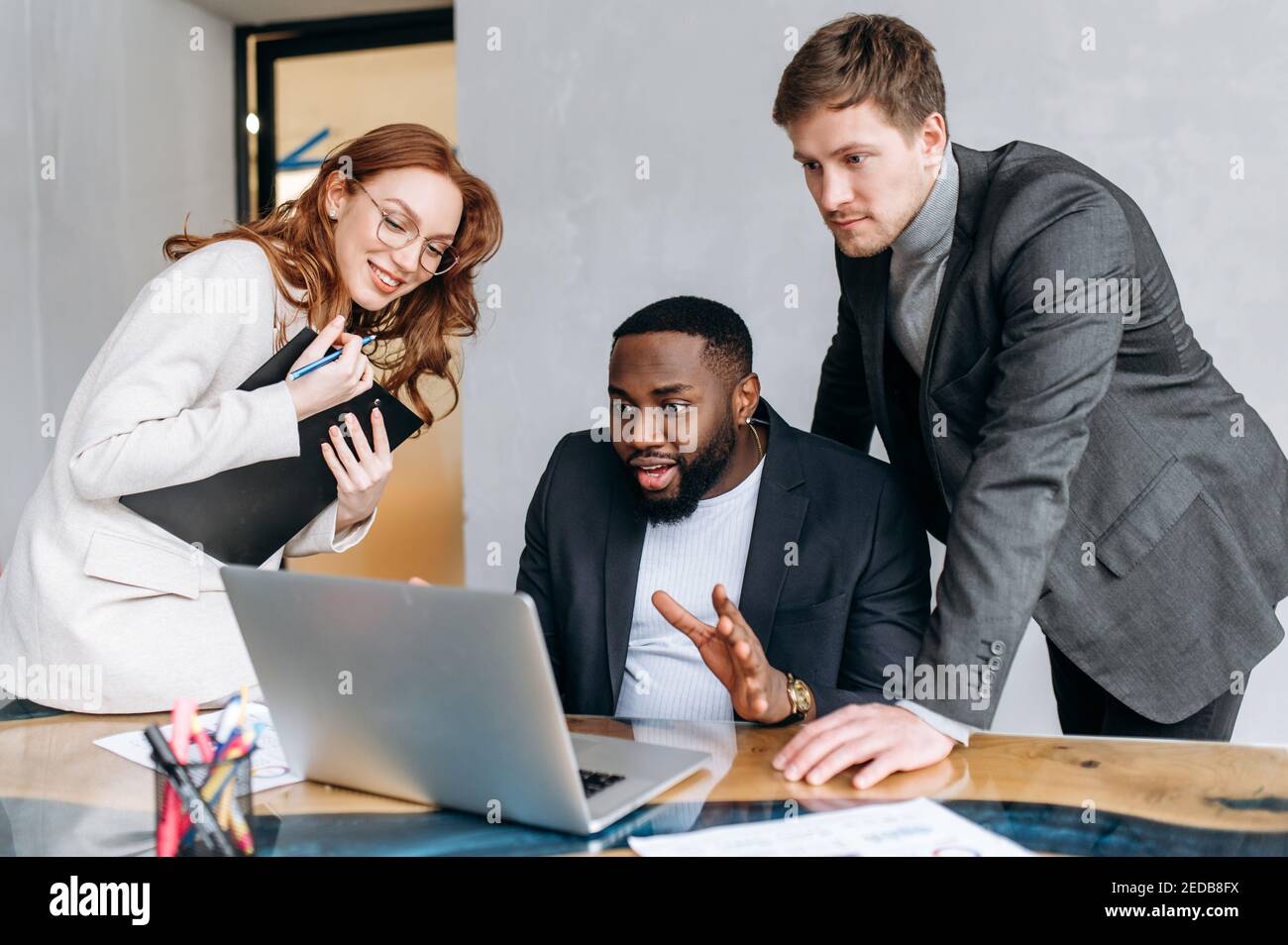 Glückliche multirassische Geschäftsleute im Büro mit einem Laptop, Blick auf den Bildschirm. Erfolgreiche Mitarbeiter lesen gute Nachrichten, erhalten Zustimmung für ein Projekt. Junge Erwachsene Mitarbeiter arbeiten zusammen und lächeln Stockfoto