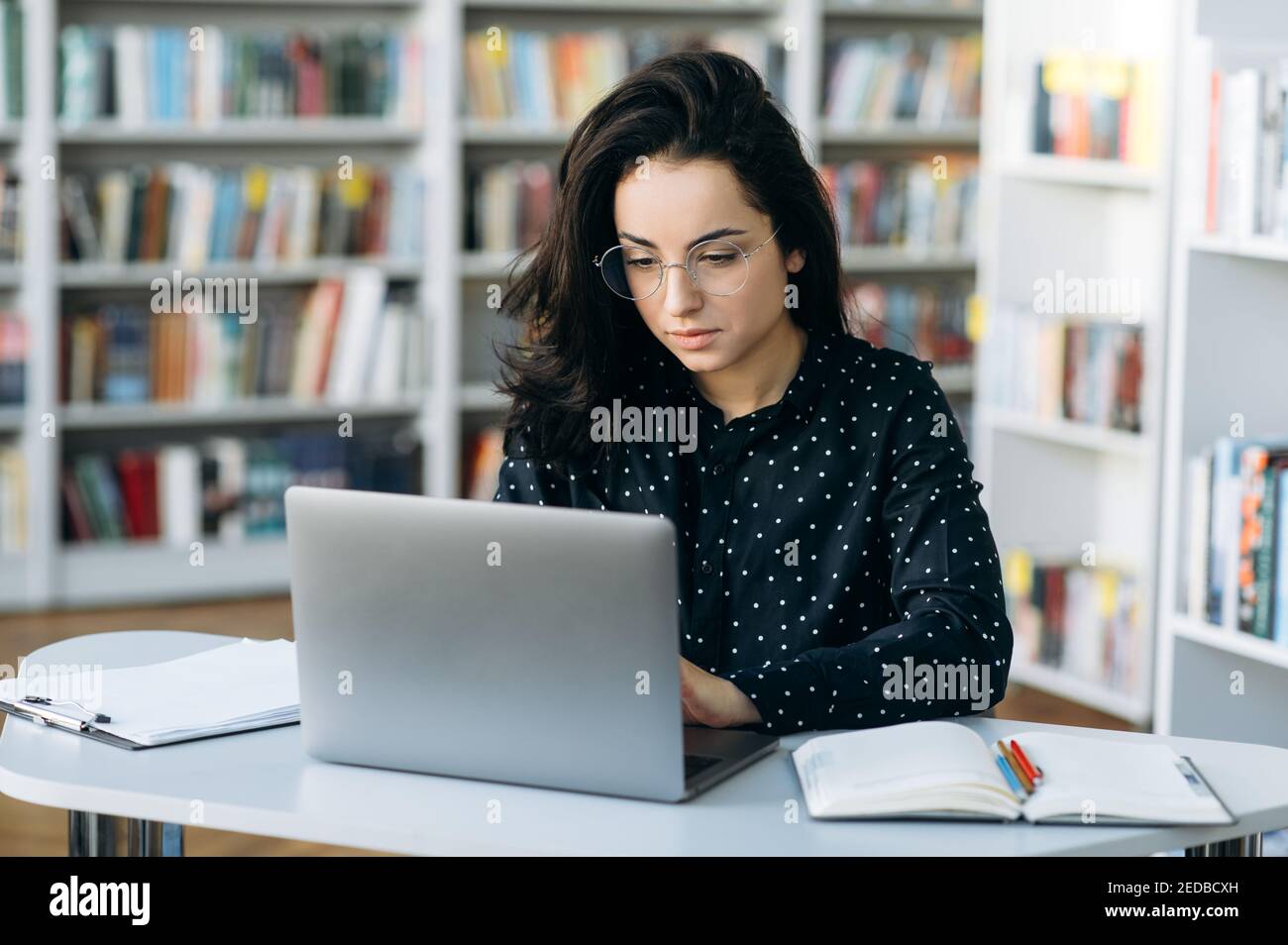 Fokussierte kaukasische junge Frau sitzen am Arbeitsplatz im modernen Büro mit Augenweide, mit Laptop. Selbstbewusste weibliche Mitarbeiterin, die eine Geschäftsstrategie plant oder mit Kollegen chattet. Stockfoto