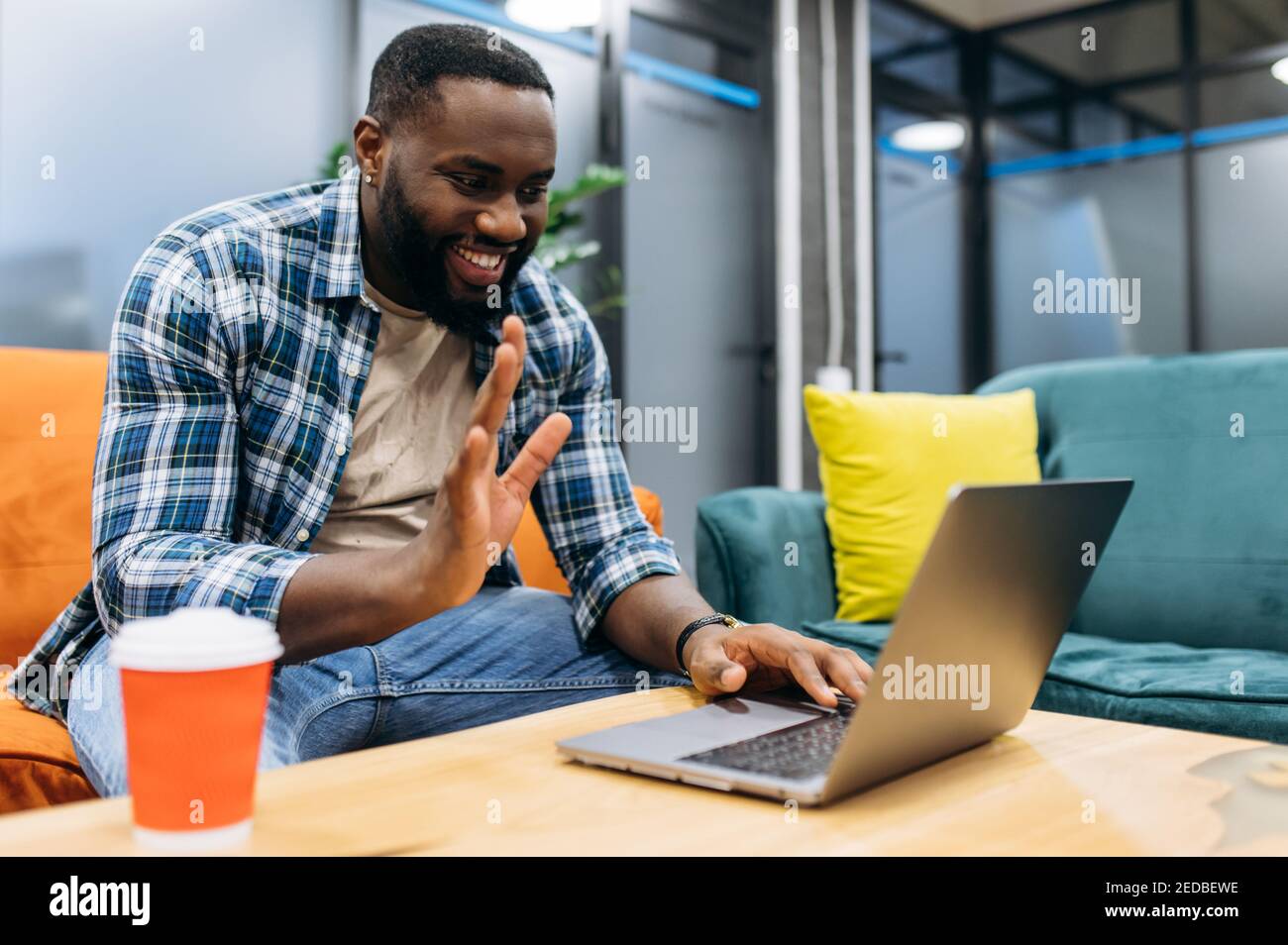 Afroamerikanischer Mann sitzt im modernen Büro in lässiger Kleidung und mit Laptop. Junge Erwachsene Geschäftsmann kommuniziert durch einen Videoanruf mit Kollegen oder Freunden, lächelnd, entfernten Arbeitskonzept Stockfoto