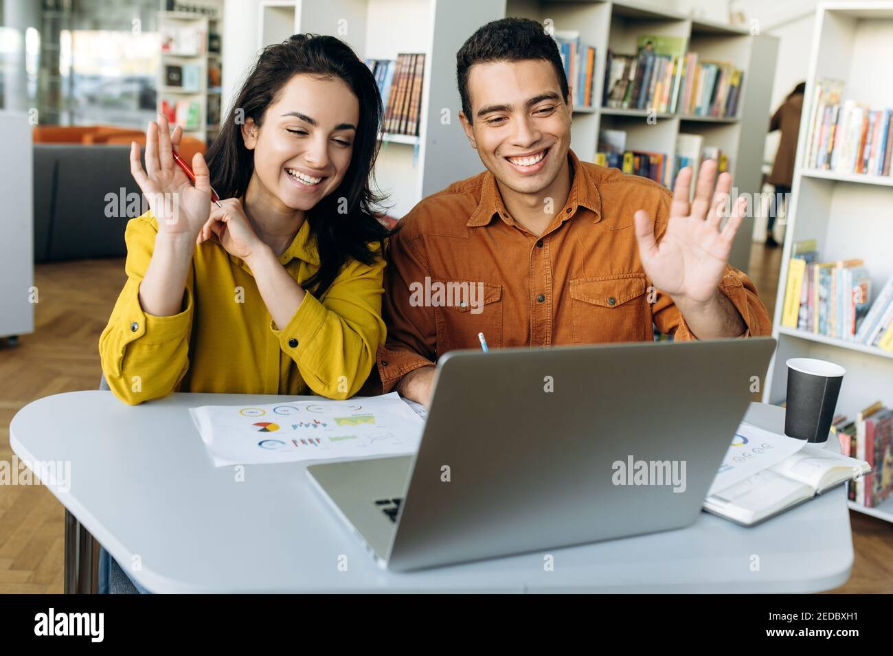 Zufriedene Kollegen oder Studenten aus verschiedenen Rassen, die am Arbeitsplatz sitzen, per Videoanruf mit Kollegen oder Freunden lächeln und auf die Laptop-Kamera blicken und die Hände winken Stockfoto
