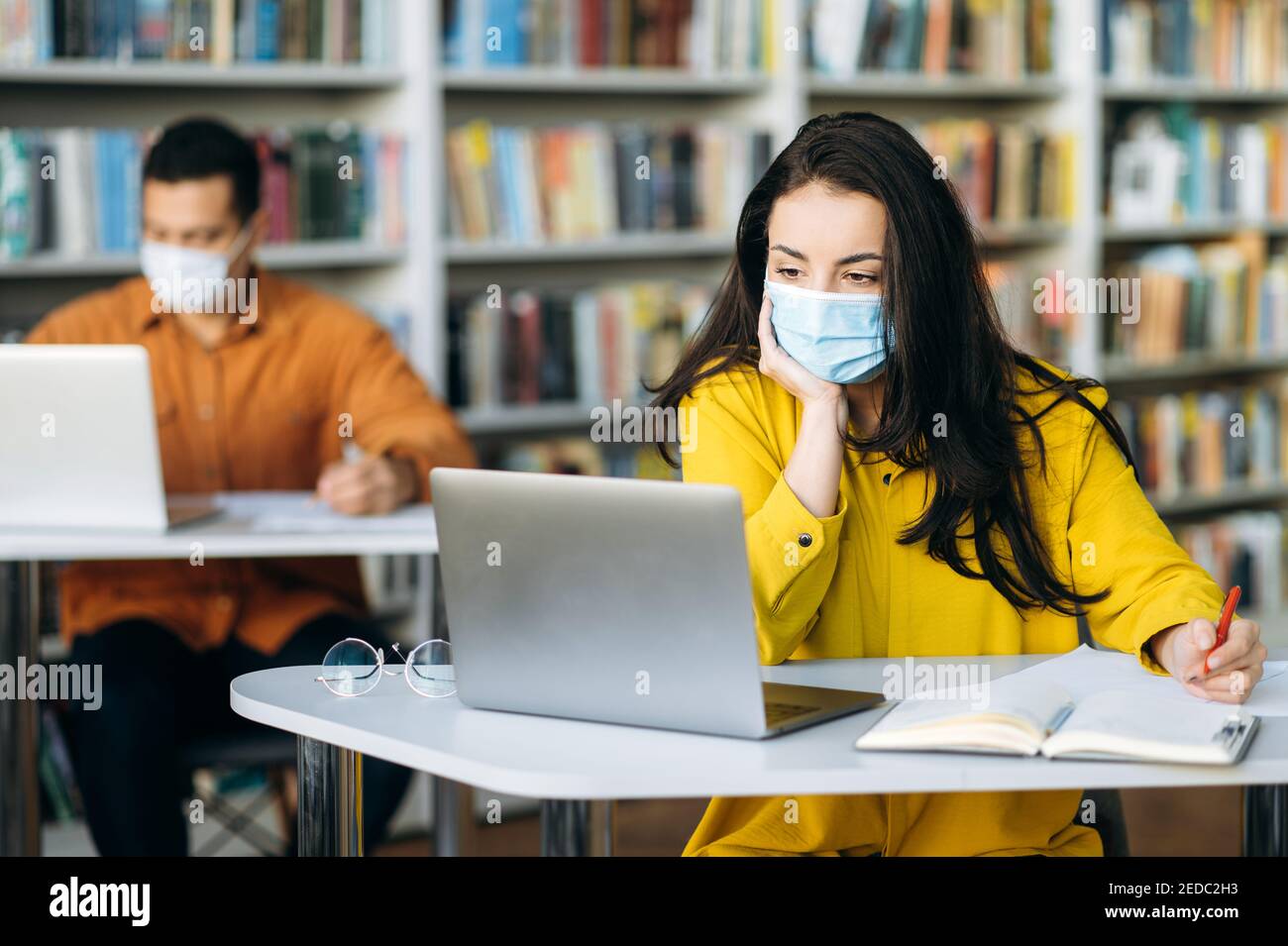 Lernen während einer Pandemie. Die Schüler sitzen in einem Abstand voneinander mit Schutzmasken auf ihren Gesichtern. Eine Schülerin sitzt während einer Unterrichtsstunde in einer medizinischen Maske an einem Schreibtisch und schaut weg Stockfoto