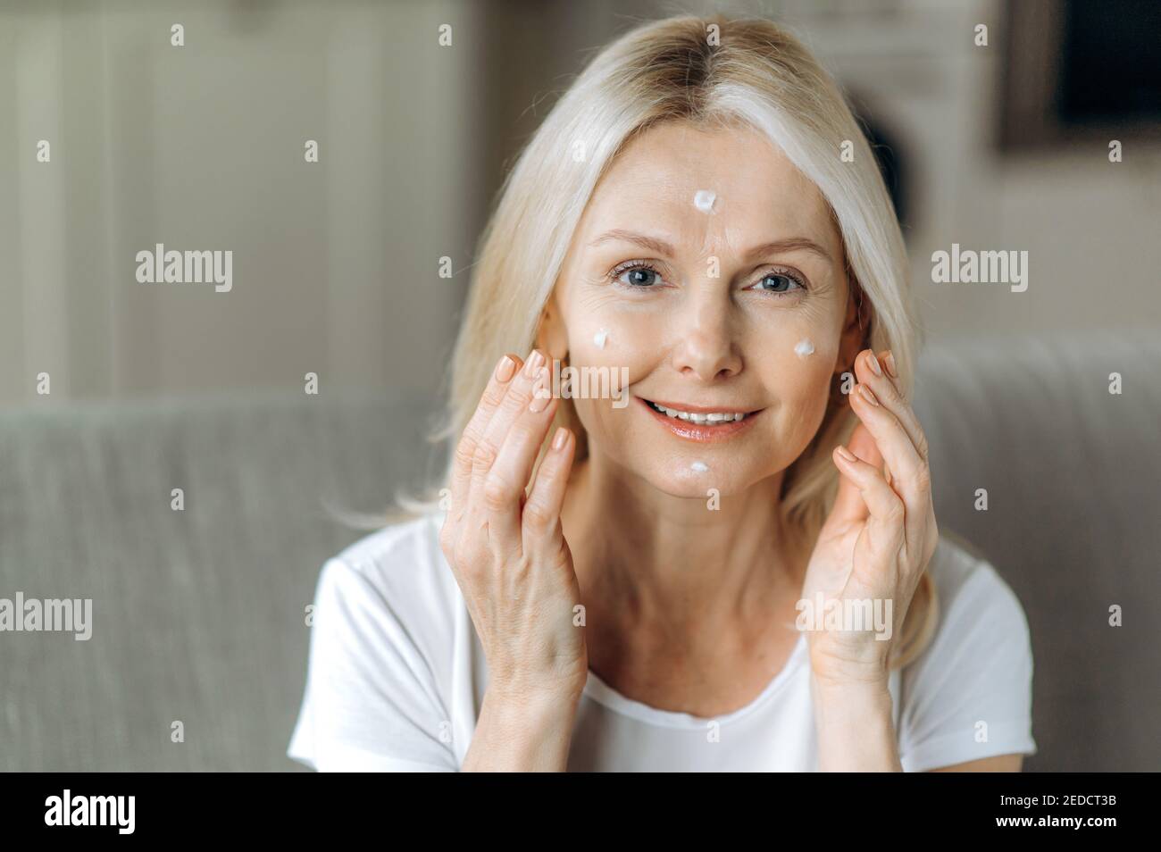 Pflege der alternden Haut. Attraktive Kaukasierin mittleren Alters kümmert sich um ihr Gesicht, wendet Anti-Aging-Faltencreme an, schaut auf die Kamera und lächelt. Nahaufnahme Stockfoto