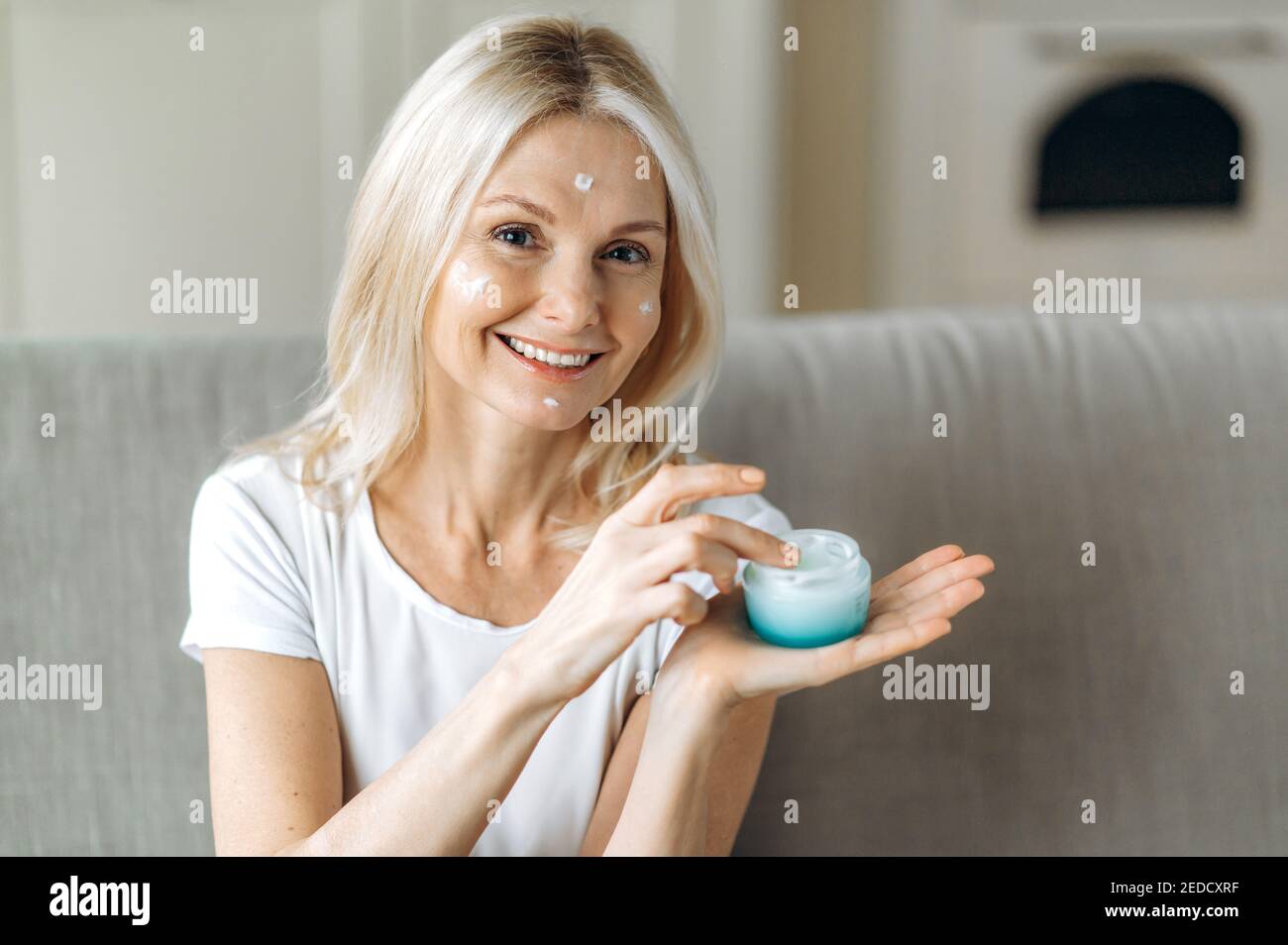 Attraktive Dame mittleren Alters kaukasischen hält Gesichtscreme in der Hand. Frau kümmert sich um ihr Gesicht, trägt Anti-Aging-Faltencreme, schaut auf die Kamera und lächelt Stockfoto