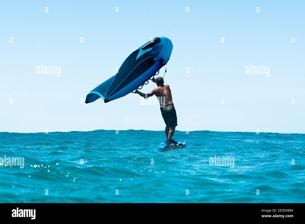 Ein Mann wingfoils mit einem Tragflügelboot Surfbrett und Hand gehalten aufblasbaren Flügel, auf einem türkisfarbenen Meer mit einem hellen blauen Himmel. Stockfoto