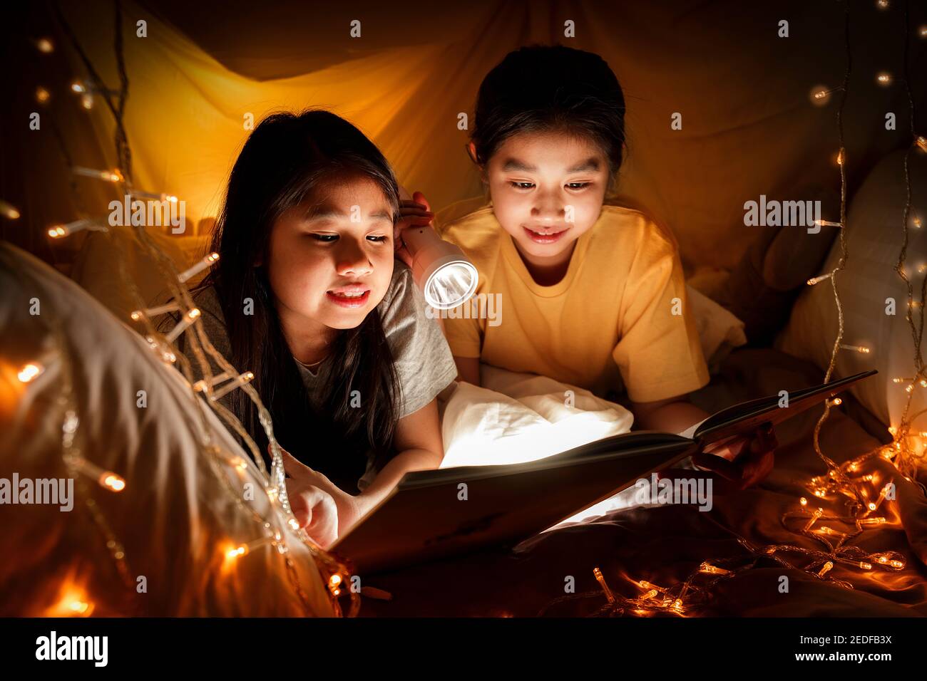 Familienkonzept. Ältere Schwester und Schwester lesen Buch mit Taschenlampe zusammen vor dem Schlafengehen. Schwester lesen gemeinsam im Bettlaken-Zelt das Geschichtenbuch. Fokus Stockfoto