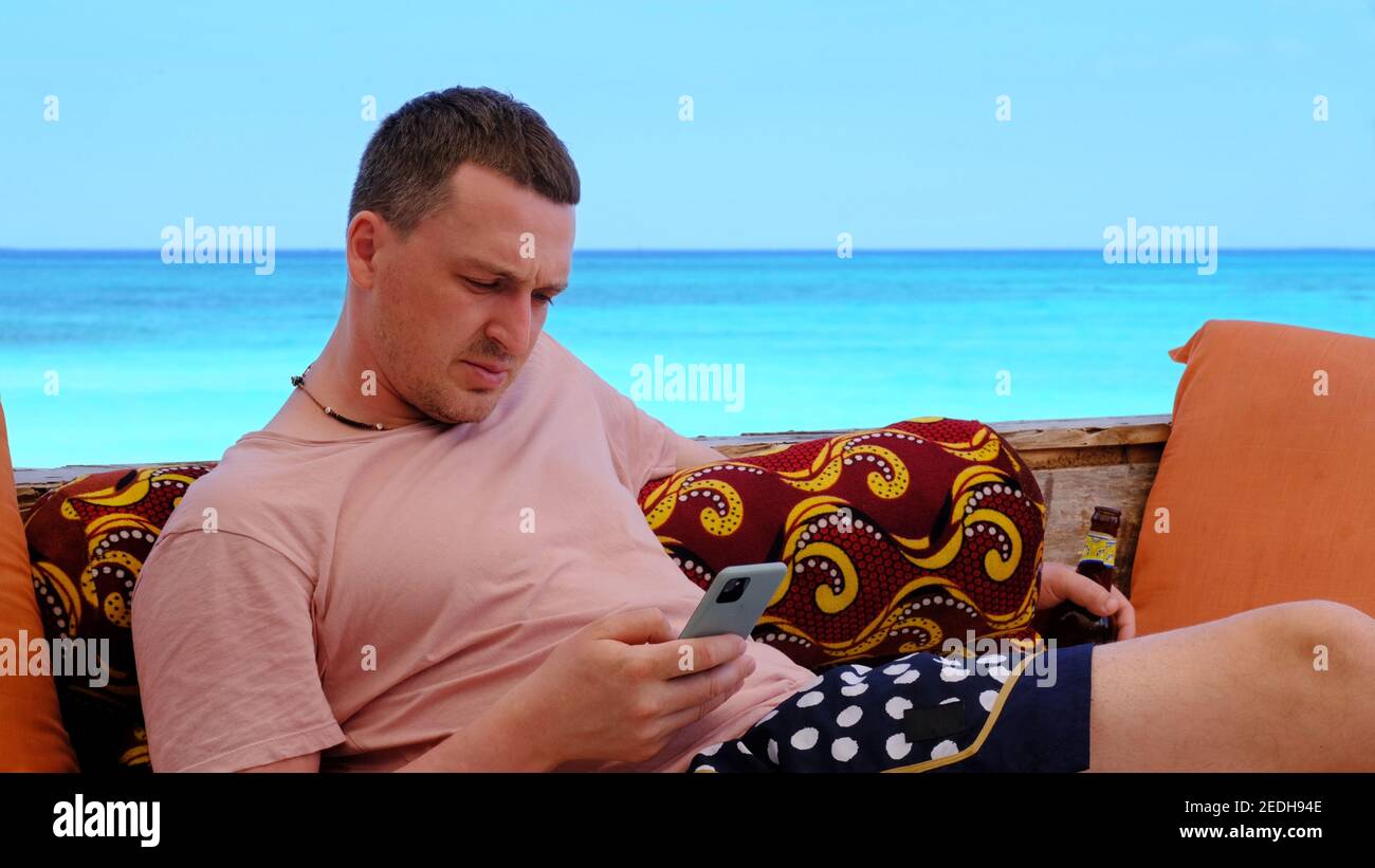 Junger Mann mit Bierflasche surft im Internet mit Telefon Komfortable Lounge-Zone am malerischen Meeresstrand im Urlaub Sonniger Tag Stockfoto