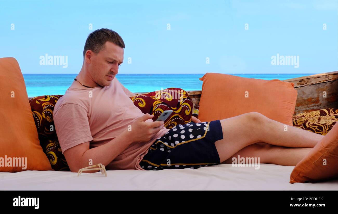 Junger Mann mit Bierflasche surft im Internet mit Telefon Komfortable Lounge-Zone am malerischen Meeresstrand im Urlaub Sonniger Tag Stockfoto