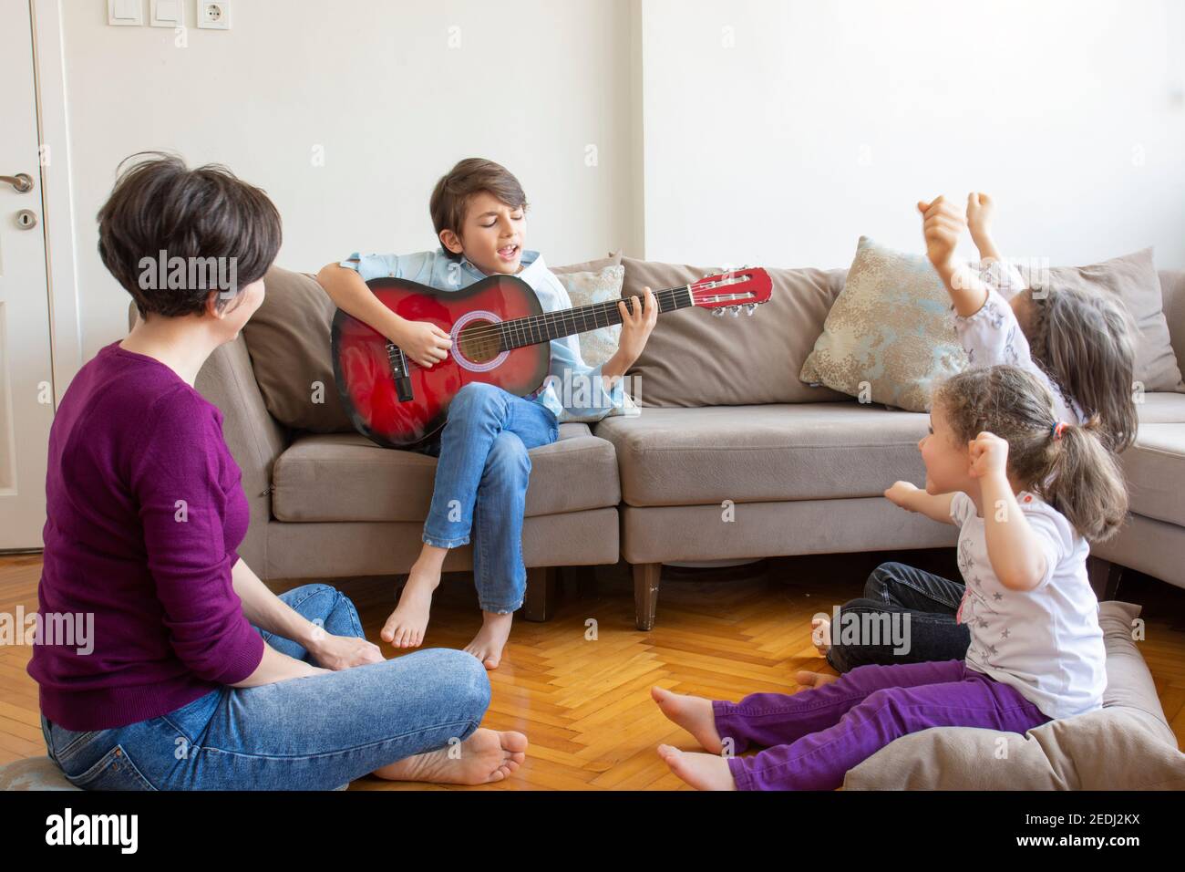 Kleiner hübscher Junge, der Gitarre spielt. Hübscher kleiner Junge, der Gitarre spielt, um sein Publikum zu Hause. Um seine Mutter, Schwester und Cousine. Stockfoto