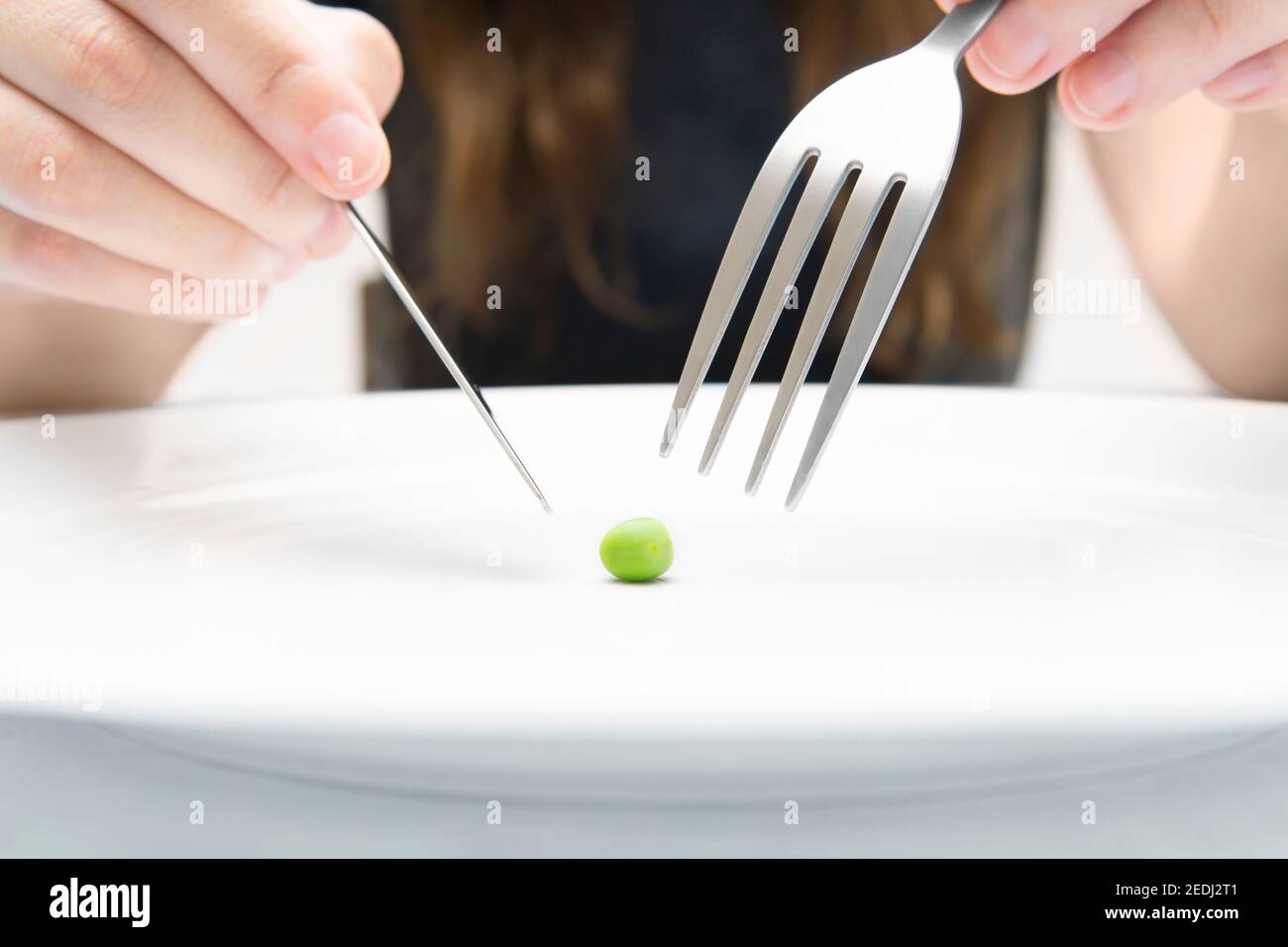Frau, die eine kleine grüne Erbse mit Messer und Gabel isst. Extreme Diät-Konzept. Nahaufnahme, selektiver Fokus. Stockfoto