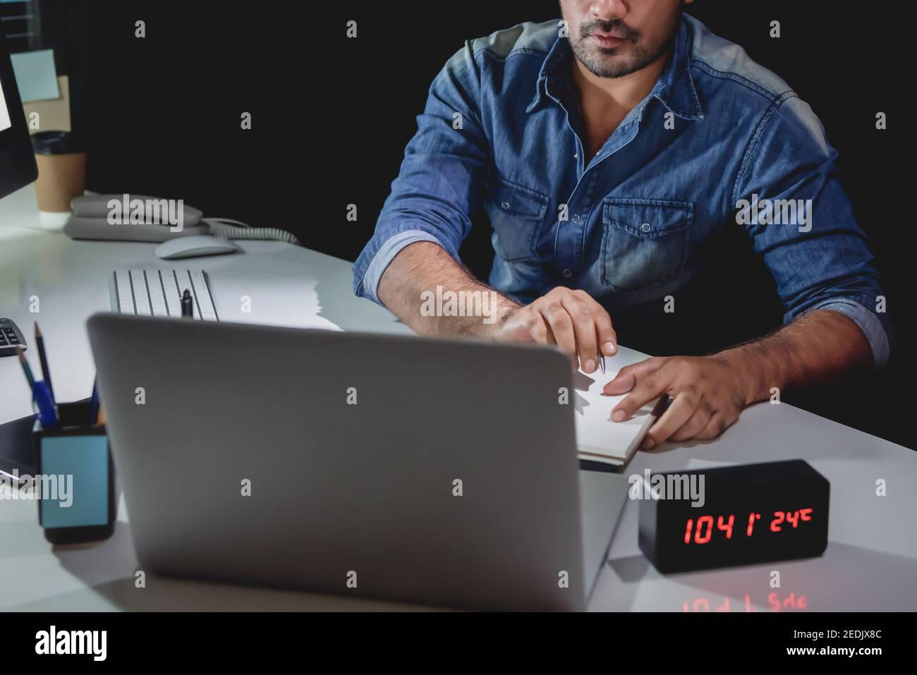 Workaholic Mann, der Überstunden spät in der Nacht im Büro bleibt Arbeiten an seinem Schreibtisch und Blick auf Laptop-Computer Stockfoto