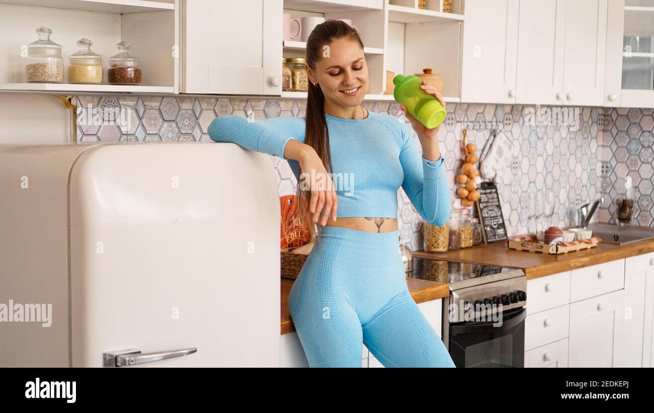 Gesundheits- und Diät-Konzept. Schöne Frau trinkt Wasser aus einer Sportflasche nach einem Training zu Hause. Sie ist in der Küche neben dem Kühlschrank. Stockfoto