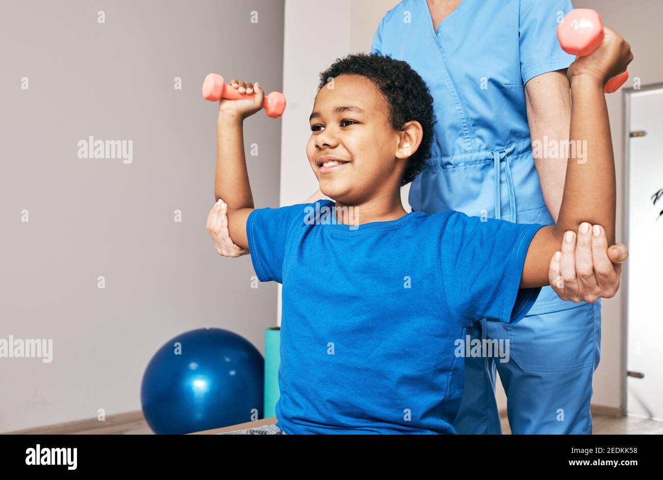 Afroamerikanischer Junge trainiert mit Physiotherapeuten mit Kurzhanteln im Reha-Zentrum. Lächelndes Kind mit den Händen nach oben, mit Hanteln. Stockfoto