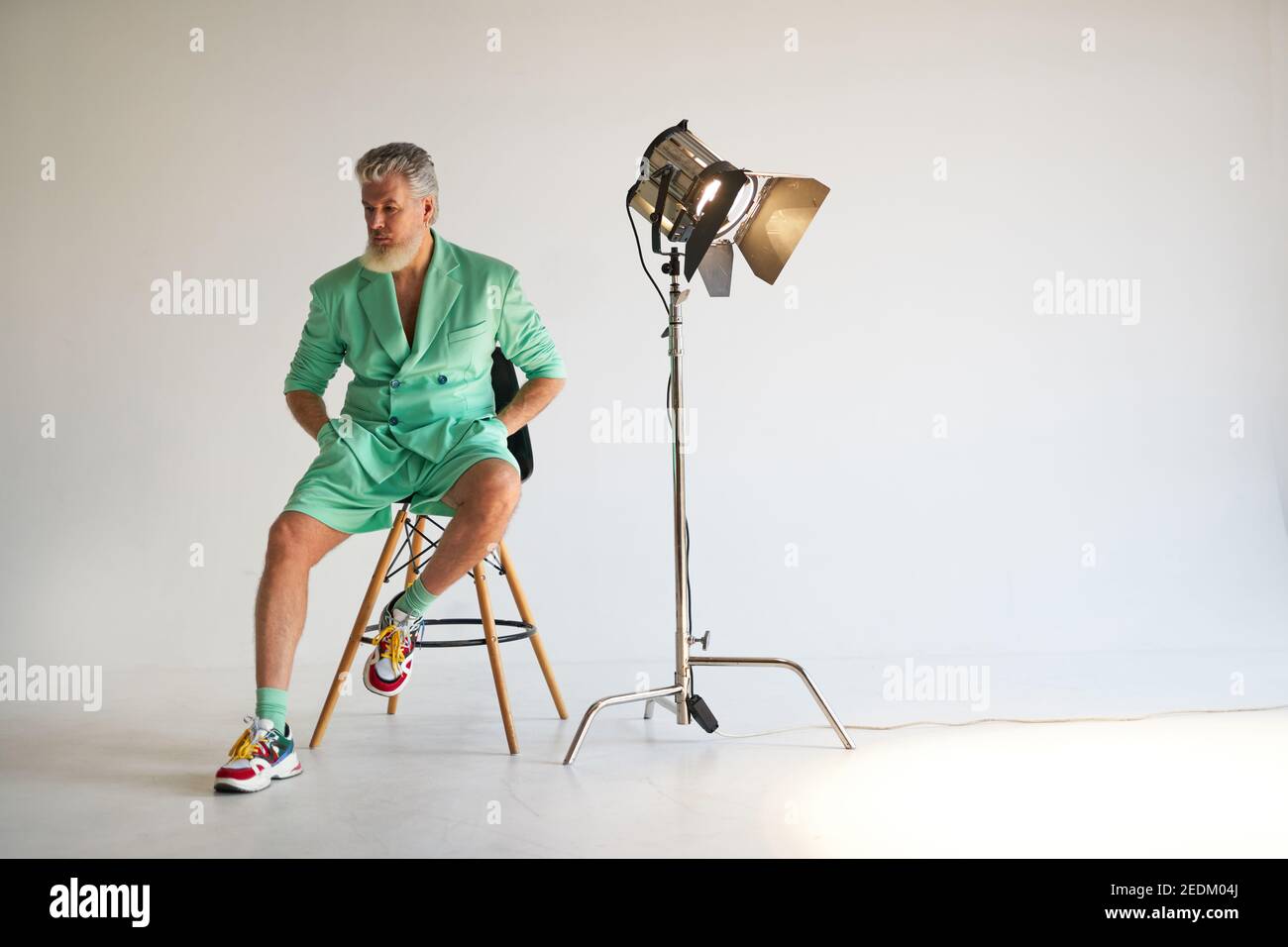 Studio-Aufnahme von trendigen grauhaarigen Mann mittleren Alters trägt stilvolles Outfit mit bunten Sneakers zur Seite schauen, sitzen neben Studio-Scheinwerfer, posiert auf weißem Hintergrund. Mode, Stilkonzept Stockfoto