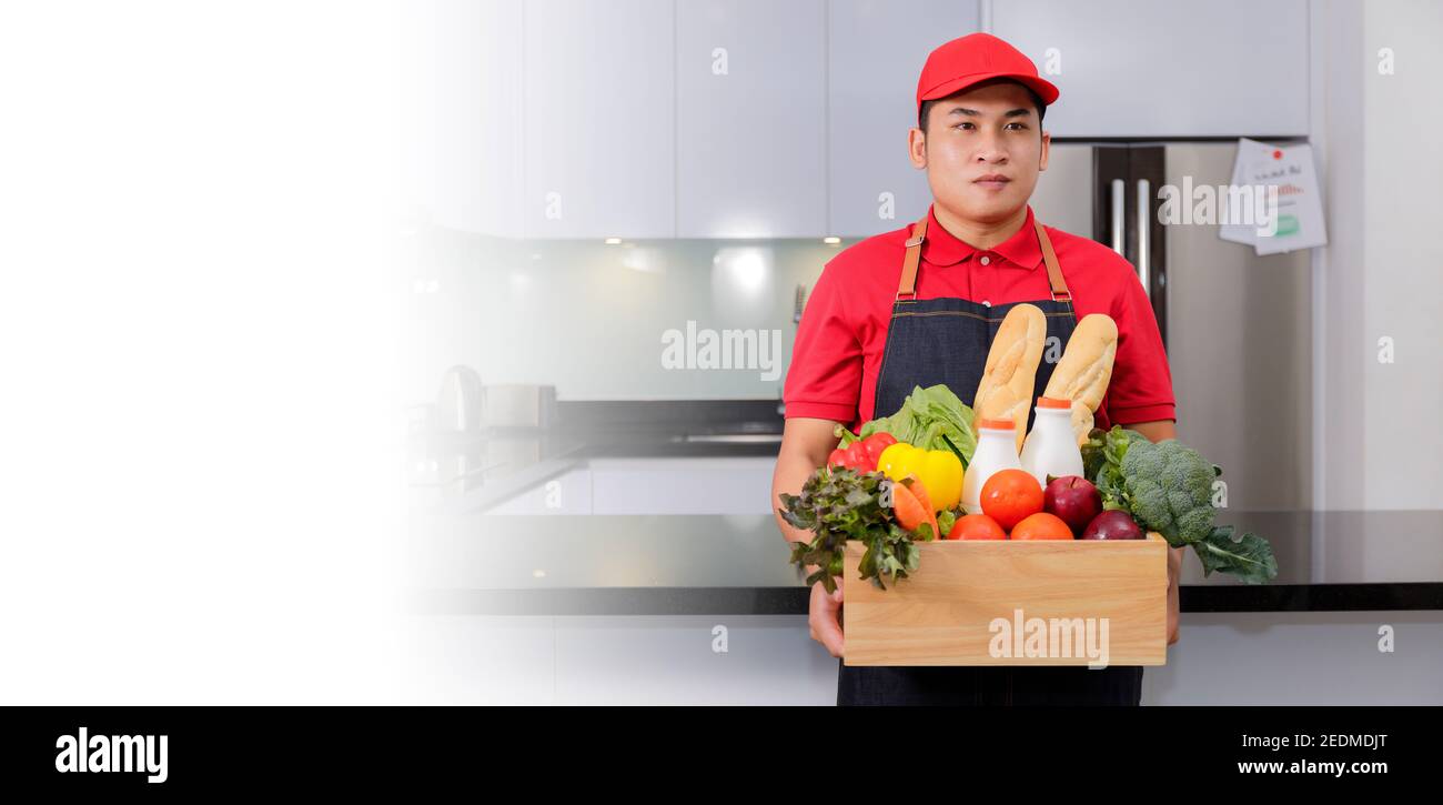 Lebensmittelgeschäft Lieferung Kurier Mann in roter Uniform und Schürze mit Lebensmittelkarton mit Lebensmitteln, frischem Obst und Gemüse. Liefert den Mann, der mit einer Holzkiste Lebensmittel umgeht Stockfoto