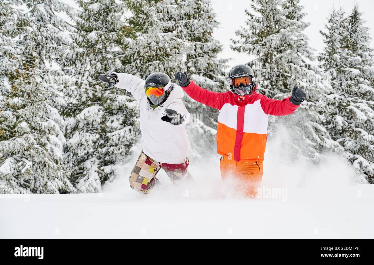 Paar Skifahrer in Skijacken und Helmen laufen durch Pulverschnee und heben die Hände, gute Zeit im Winterwald mit verschneiten Bäumen auf dem Hintergrund. Konzept von Freizeit, Spaß und Beziehungen. Stockfoto