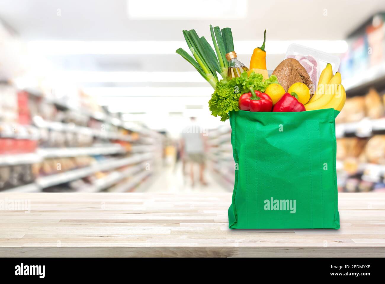 Lebensmittel und Lebensmittel im grünen umweltfreundlichen wiederverwendbaren Einkaufstasche auf Holztisch mit verschwommenem Vormarkt Gang im Hintergrund Stockfoto