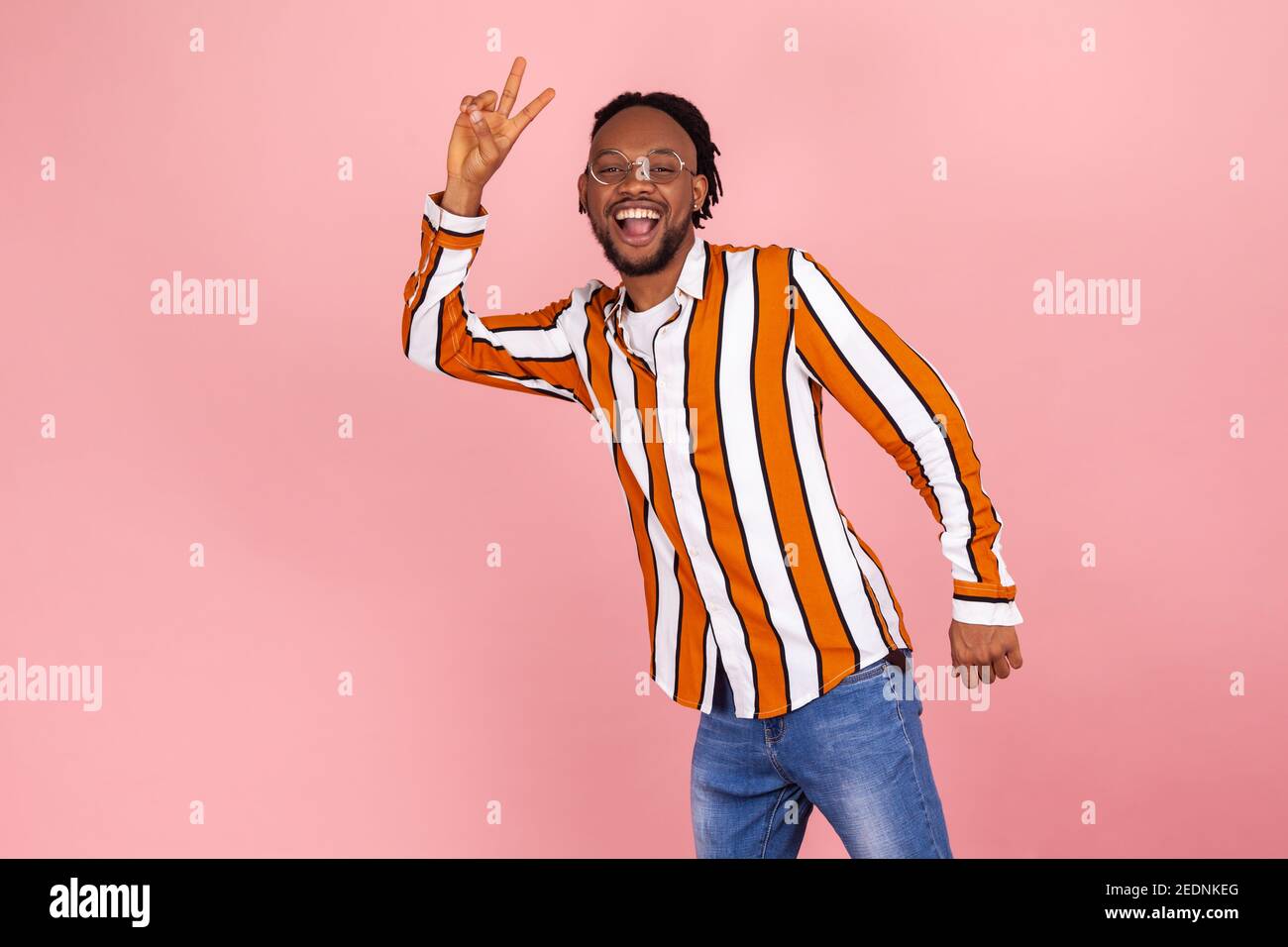 Freundlich lächelnder afrikanischer Mann mit Dreadlocks im trendigen gestreiften Hemd und Denim-Hose mit Siegesgeste mit Fingern und Blick auf die Kamera. Innen s Stockfoto