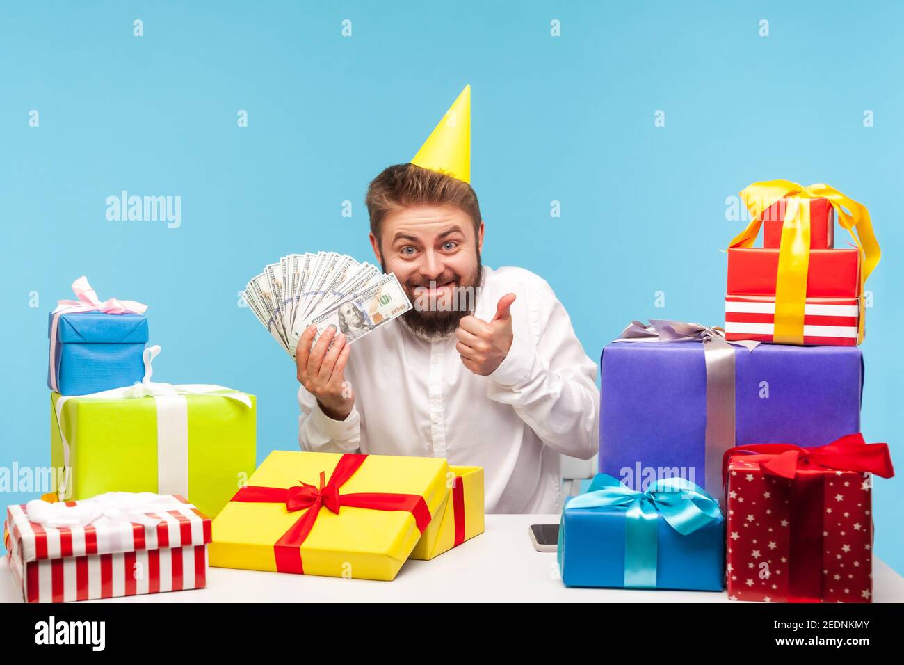 Aufgeregt bärtigen Mann in Kegelhut Eröffnung Geschenk-Box und zeigt Daumen nach oben viel Dollar-Bargeld erhalten, zufrieden mit Geld Bonus, Gehaltserhöhung. Indo Stockfoto