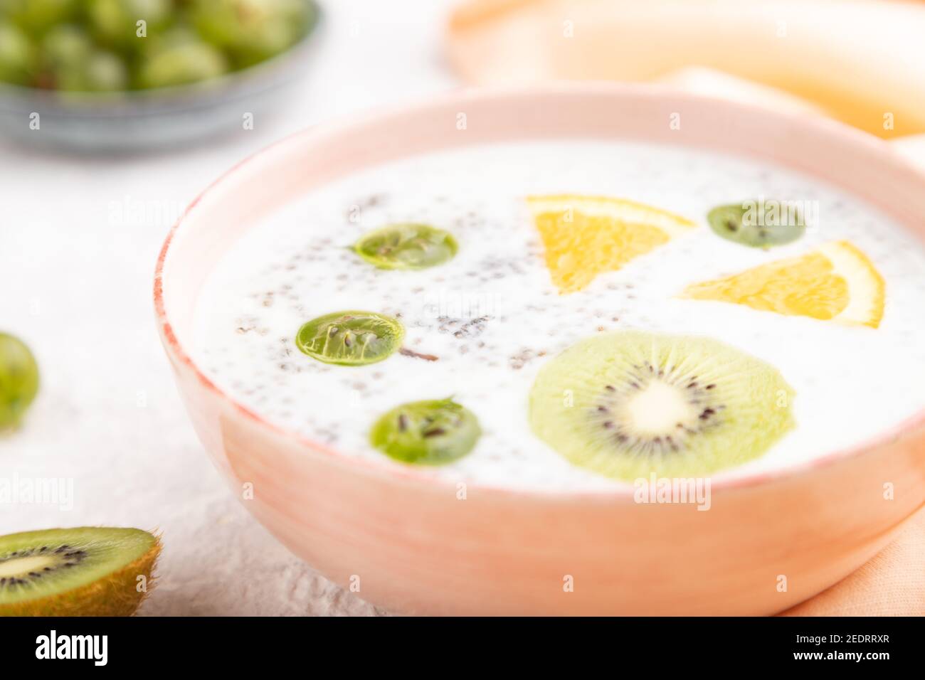 Joghurt mit Kiwi, Stachelbeere, Chia in Keramikschale auf grauem Betongrund und orangefarbenem Leinentextilien. Seitenansicht, Nahaufnahme, selektiver Fokus. Stockfoto