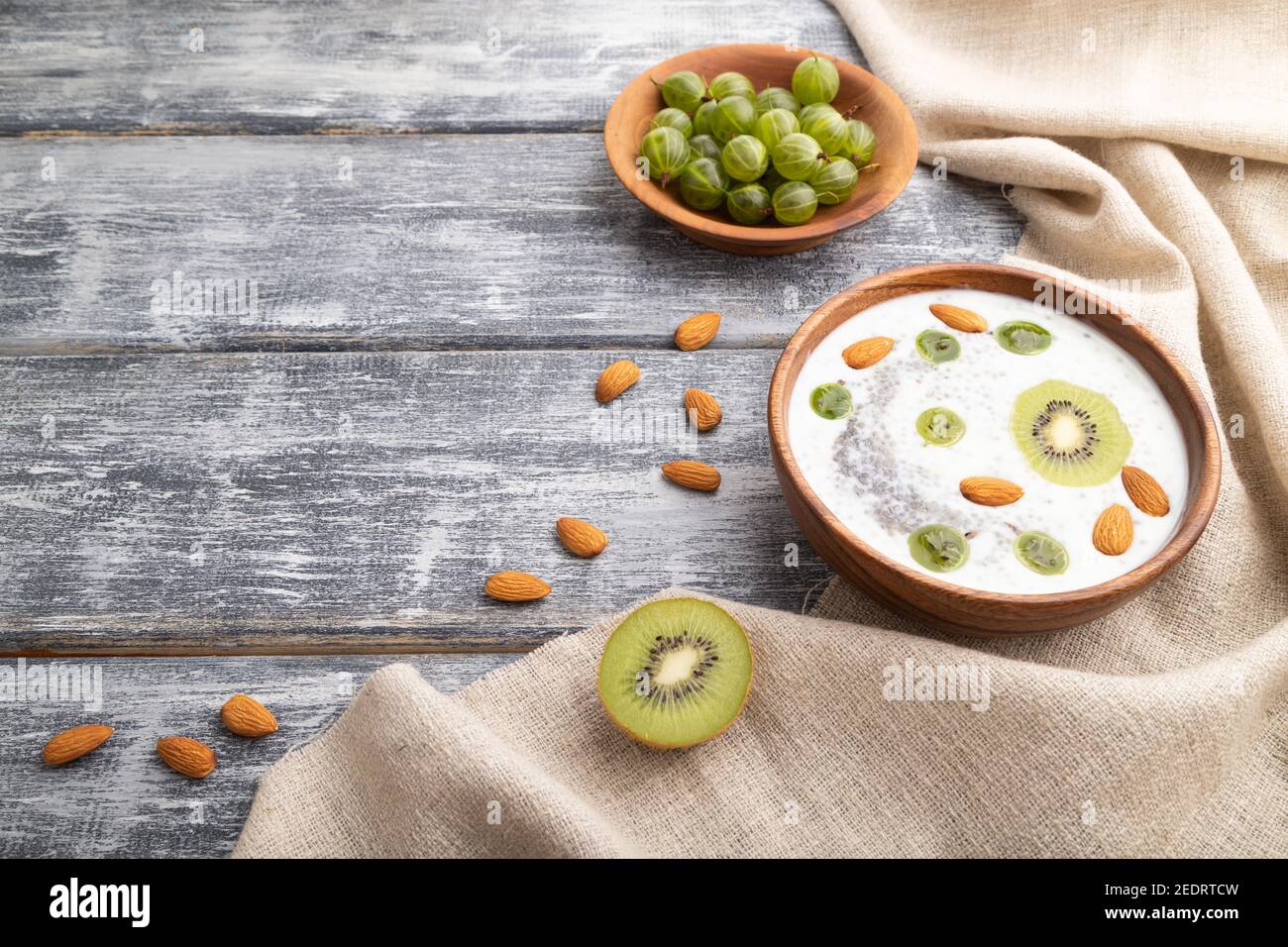Joghurt mit Kiwi, Stachelbeere, Chia und Mandeln in Holzschüssel auf grauem Holzhintergrund und Leinentextilien. Seitenansicht, Kopierbereich. Stockfoto